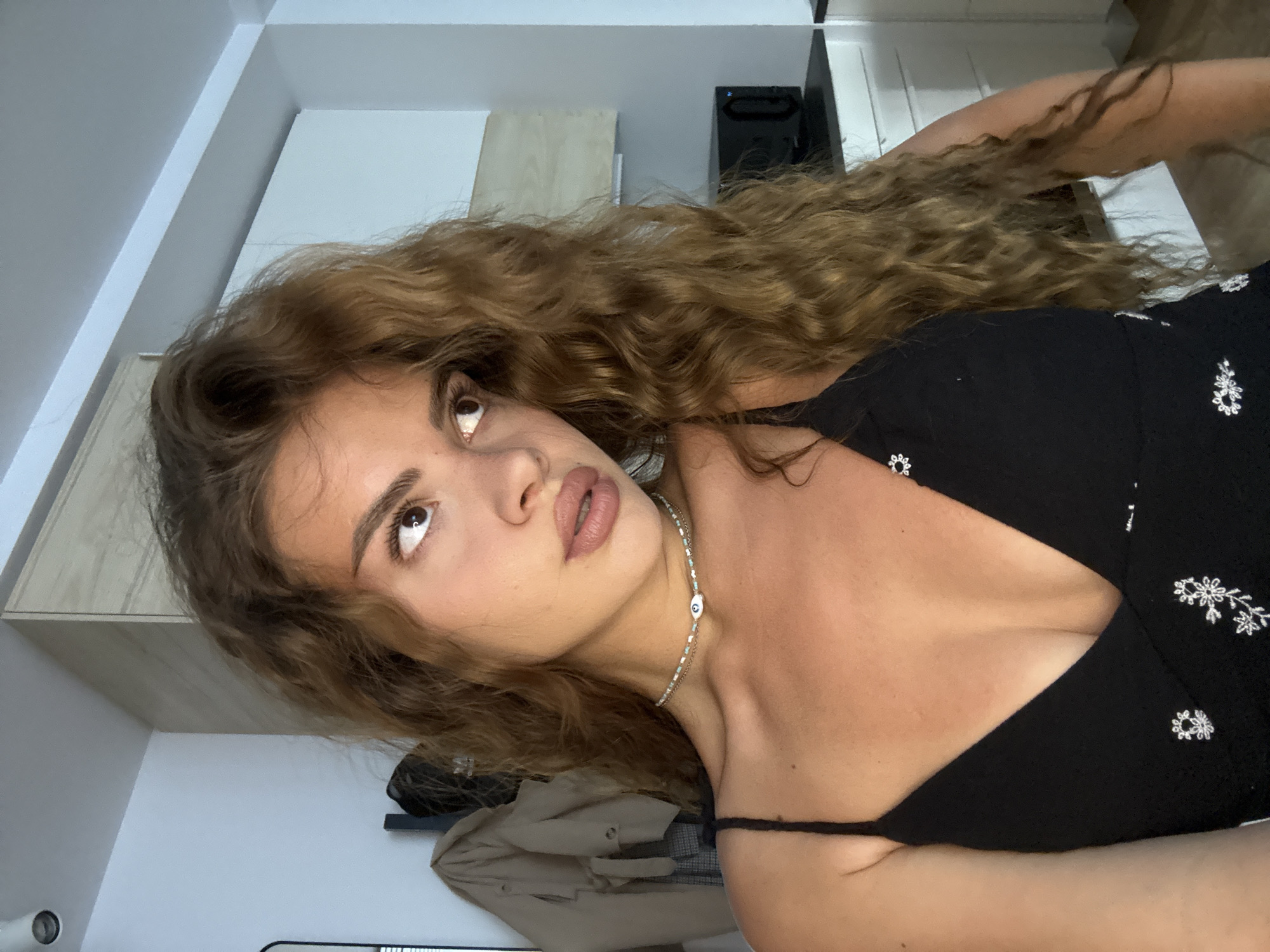 A woman with brown, curly hair is photographed indoors. She is wearing a black floral-patterned top and a necklace with a white and blue bead. The background shows a white wall with cabinets and what appears to be a dark speaker or electronic device. A beige jacket or coat is visible near the bottom of the frame. The location is unknown.