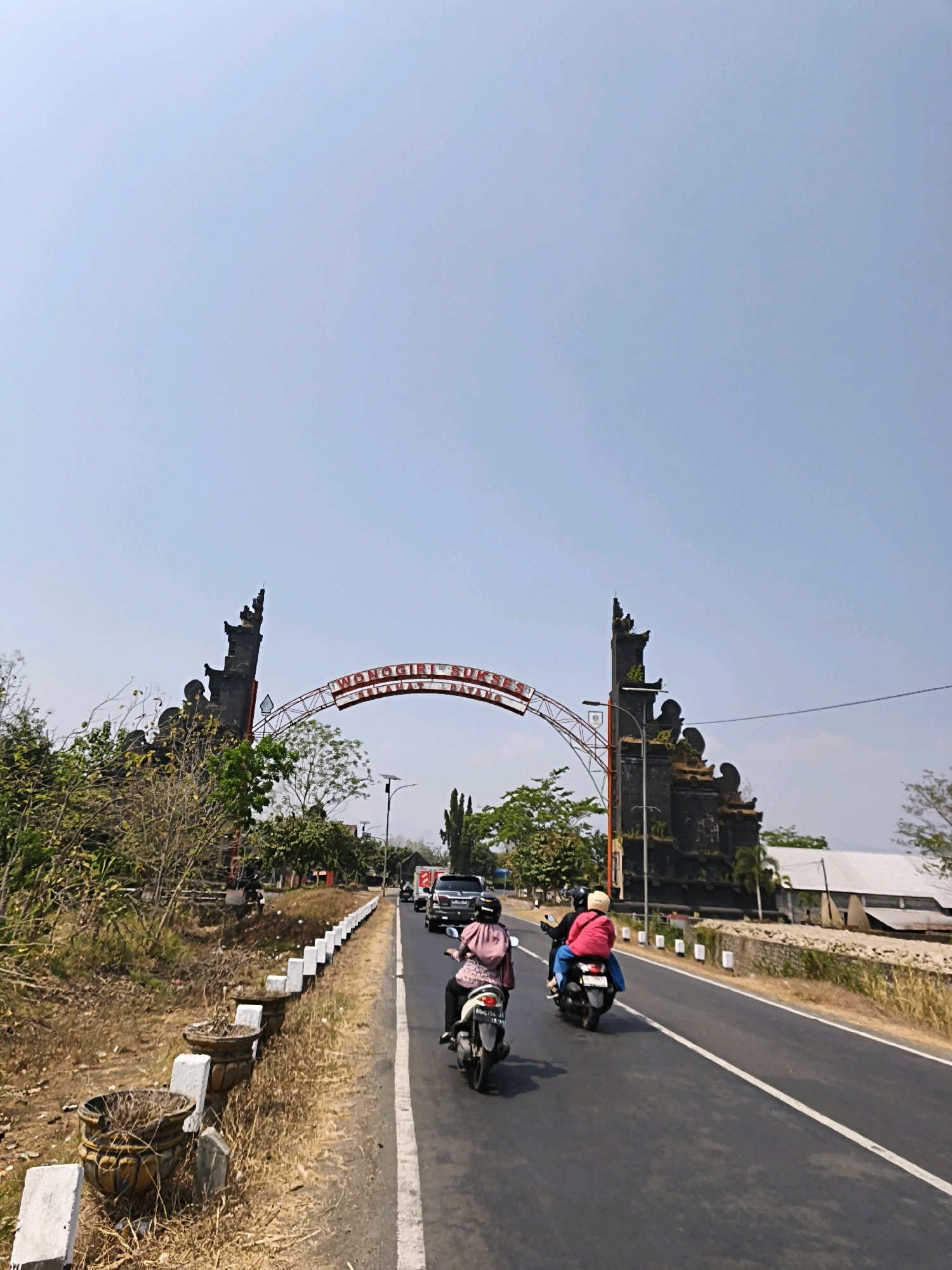 In the city of Nambangan, Indonesia, a road leads towards an ornamental archway with the inscription "WONOGIRI SUKSES SELAMAT DATANG." The arch is supported by two dark, intricately carved pillars. The sky is a pale blue with no clouds. On the right side of the road, two people are riding on a single motorcycle, with the passenger wearing a pink jacket and a white helmet, and the driver wearing a pink shirt and a black helmet. On the left side of the road, a dry, grassy embankment is lined with decorative concrete planters. The road is asphalt with a white dividing line. Further down the road, several cars and motorcycles are visible. The overall environment suggests a warm, sunny day.