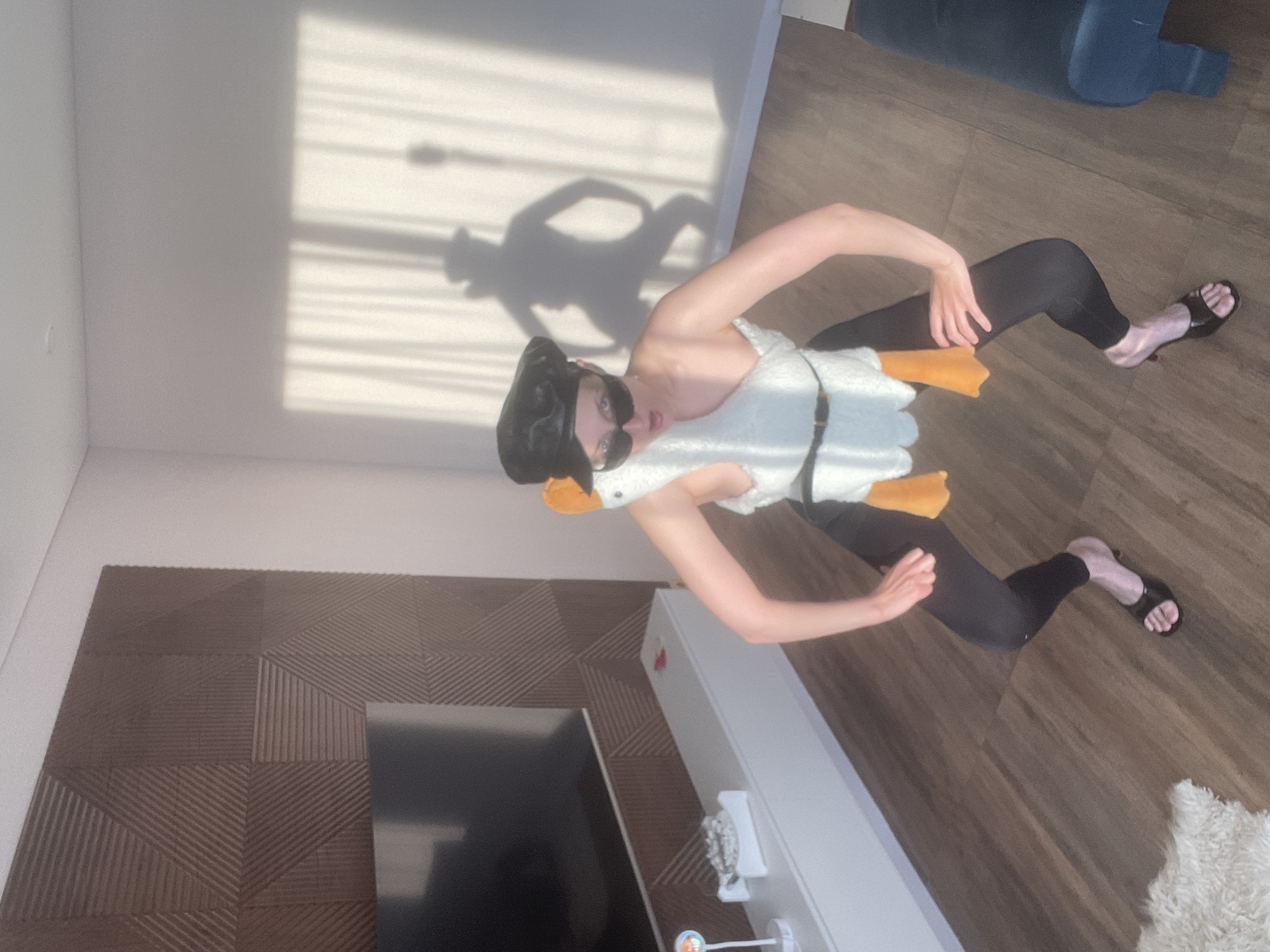In a brightly lit room in Podshivalovo, Russia, a person stands posing humorously in a goose costume. The individual wears a black beret-like hat, dark sunglasses, and a white, fluffy goose-shaped top featuring orange beak and webbed feet details. This is paired with black leggings and black open-toed sandals. The person’s body is slightly twisted, and they gaze directly at the camera, suggesting a playful or comedic intent, possibly for a costume event or photo.
The setting is a modern interior with light wooden flooring and clean white walls. A decorative textured wooden panel covers a section of the wall in the lower left, behind a flat-screen television set. Below the television is a white, low-profile console table adorned with small decorative items, including a stand with a round object and something metallic. A white, shaggy rug is visible on the floor in the lower right. Direct sunlight streams into the room, casting a distinct shadow of the person and the horizontal lines of window blinds onto the white wall behind them, indicating it is daytime, likely morning or afternoon. A large, dark blue or grey object, possibly furniture, is partially visible in the upper right. There is no visible text in the image.