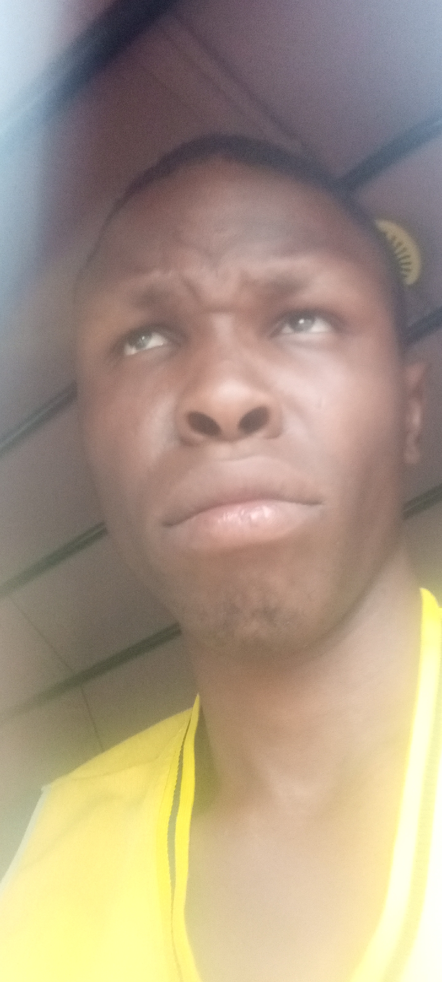 This close-up portrait features a young man with dark skin, positioned at an upward angle. He is looking upwards and slightly to his left with a furrowed brow, indicating an expression of contemplation, thought, or mild concern. His lips are slightly pouted. He is wearing a bright yellow garment, possibly a t-shirt or jersey, with a visible black trim detail on the shoulder/neckline. The background is indistinct and appears to be an indoor or enclosed setting, possibly a vehicle interior, with faint linear patterns visible on what could be a ceiling or upper wall. There is a noticeable glare or overexposure effect in the upper left portion of the image. No text is visible. The scene is set in Sabon Tasha, Nigeria.