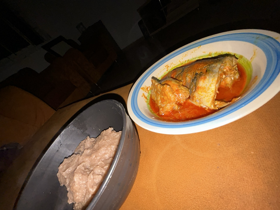 The scene depicts an indoor dining setting in Egbe, Nigeria, under dim lighting conditions. Two bowls of food are positioned on a textured, reddish-orange surface. In the upper left, a black bowl contains a substantial mound of light brown, semi-solid food with a smooth appearance. In the lower right, an oval-shaped white bowl, detailed with blue and light green stripes along its rim, holds two pieces of cooked fish submerged in a vibrant reddish-orange stew. Dark, indistinct furniture is partially visible in the dimly lit background. No individuals or specific actions are present within the frame.