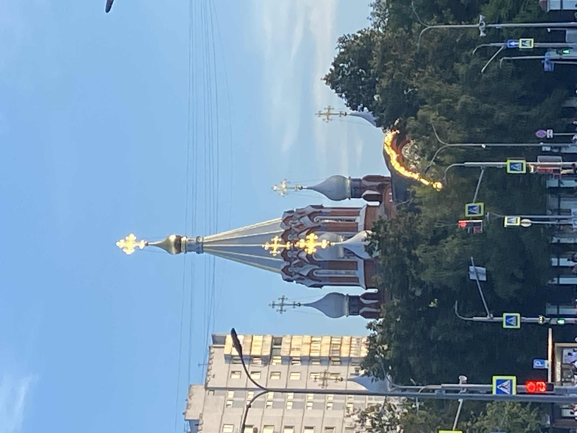 This is a vertical, medium-distance, eye-level shot taken in Moscow, Russia, capturing an Orthodox church with multiple onion domes and a tall, slender central dome topped with a golden cross. The church is predominantly red brick with white decorative elements and golden accents. To the left of the church, a tall, modern apartment building with numerous windows is visible. The scene is framed by lush green trees, and several streetlights and traffic signals are present in the foreground. The sky is a clear, bright blue. A bright yellow, curved light fixture or decoration is draped over the trees in the mid-ground, between the church and the apartment building. Pedestrian crossing signs and traffic lights indicate a busy urban intersection. The overall lighting suggests it is daytime, possibly late afternoon or early evening due to the warm glow on the church's golden elements. No people are clearly discernible in the image.