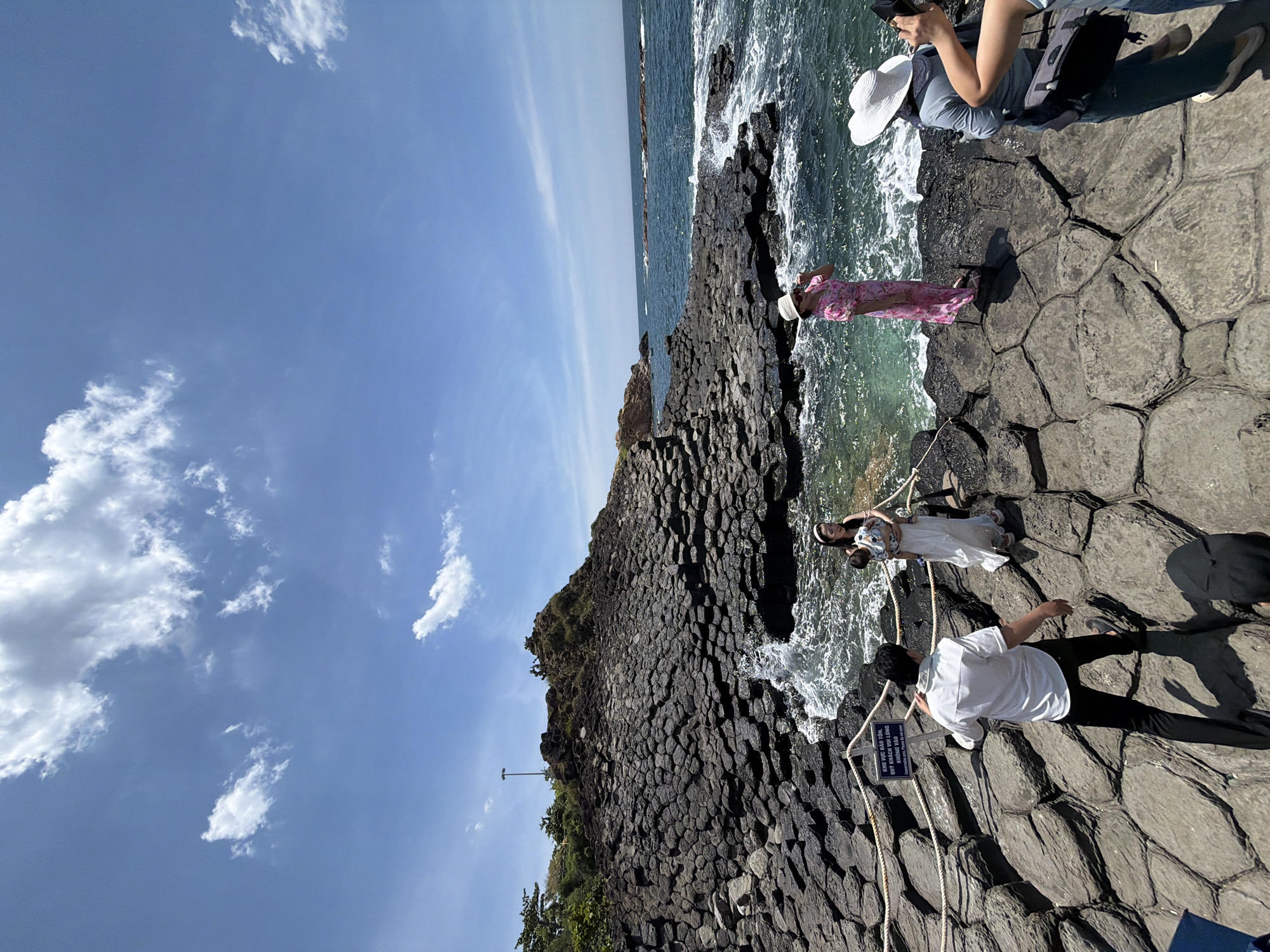 The image depicts the Gành Đá Đĩa (The Cliff of Stone Plates) in Phú Lương, Vietnam. The landscape is characterized by a dramatic formation of hexagonal basalt columns that descend into the sea. The sky is a clear blue with scattered white clouds. In the foreground, several people are present on the rocky shore. On the left, a man in a white shirt and black pants is walking away from the camera. To his right, a woman in a white dress and a man holding a baby are positioned near the water's edge. Further to the right, a woman in a pink floral dress and a white hat is facing the sea, and another person in a blue shirt and a white hat is seen from the back, holding a camera. The ocean exhibits moderate waves crashing against the rocks. A rope barrier with a sign is visible in the lower left quadrant of the image. The scene captures a tourist destination with natural geological formations and visitors engaging with the environment.