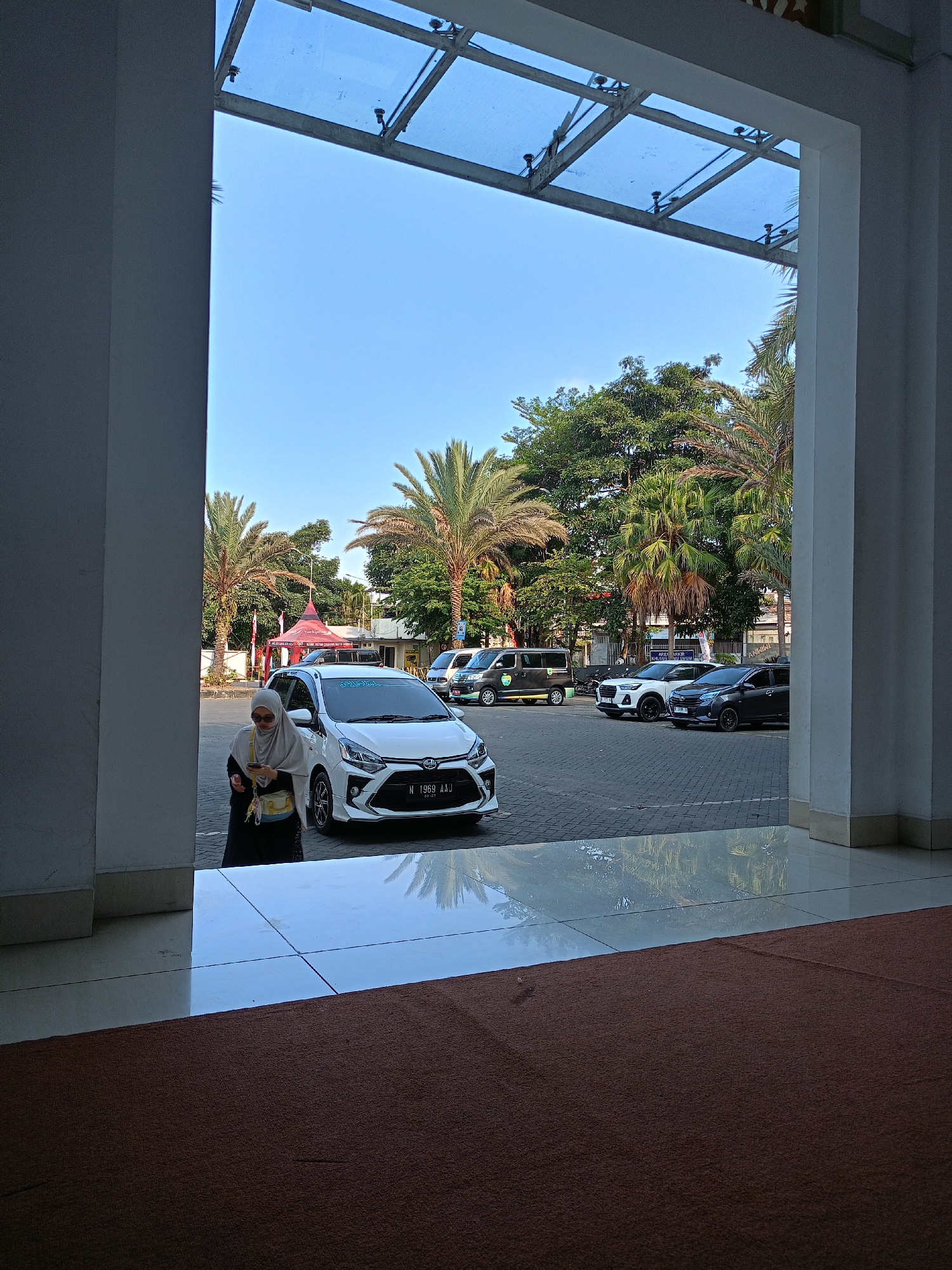 A full, eye-level, medium shot shows a view of an outdoor parking lot and trees through an open entryway. The entryway is framed by large, white, rectangular columns on the left and right. A glass-paneled roof is visible above, supported by a metal frame. The foreground is dominated by a dark brown carpet. To the left, a white car is parked, and a woman wearing a hijab and a dark dress is standing next to it, looking down at her phone. The parking lot is paved with gray interlocking stones and contains several other vehicles, including a black van, a white SUV, and a dark sedan. Palm trees and other lush green trees are scattered throughout the background. The sky is clear and blue. The scene is likely located in Batu, Indonesia, a city known for its natural beauty and tourist attractions.
