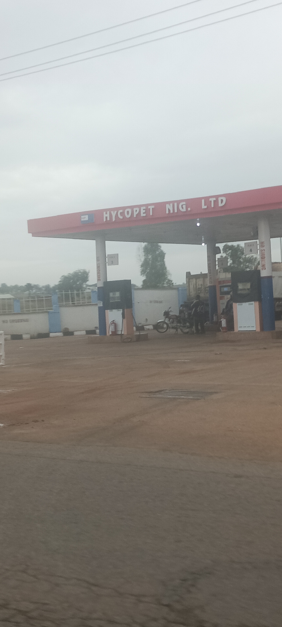 The image shows a Hycopet Nig. Ltd. gas station in Ladura Foron, Nigeria. The gas station has a red roof with white lettering displaying the company name. Several gas pumps are visible under the roof. A motorcycle is parked near one of the pumps, where people appear to be refueling. A fire extinguisher is mounted near a pump. In the background, there is a truck with a white container. The sky is overcast, and the ground appears to be dirt or gravel. Electrical power lines are visible in the upper part of the image.