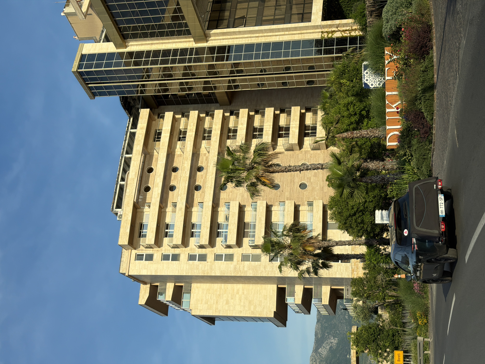 The image shows a beige, multi-story building with a glass-paneled structure on top. It features rows of windows, circular design elements, and palm trees in front. A black van is parked on the side of the road, partially obscuring a sign that reads "DUKLEY". The location appears to be in Bečići, Montenegro, based on the sign and general appearance of the buildings and landscape. The sky is clear and blue.
