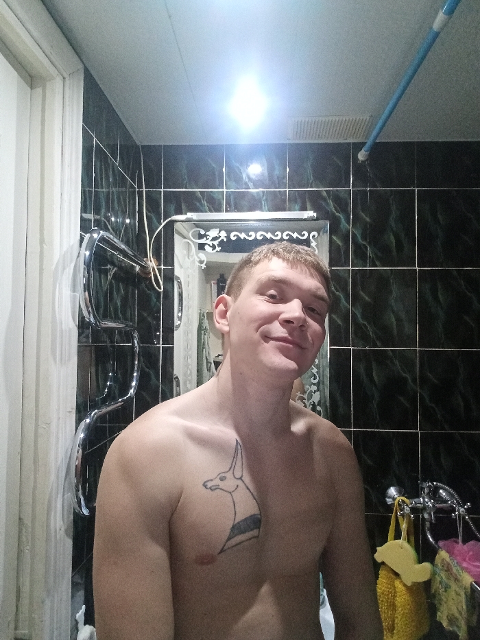 A shirtless male, appearing to be in his twenties, stands in a bathroom and smiles directly at the camera. He has short, light brown hair and a tattoo of a stylized animal head with a dark striped neck on his left pectoral muscle. The bathroom walls are covered with dark green or black rectangular tiles featuring a marbled pattern. A bright light fixture is visible on the white ceiling, emitting a glare in the upper center of the image.
To the left of the male subject, a chrome S-shaped heated towel rail is mounted on the tiled wall. To his right, behind his shoulder, a mirror with a decorative, ornate silver frame is visible, reflecting a portion of the bathroom behind the camera, including what appears to be a shower curtain or additional hanging items. Below the reflected mirror, a chrome shower/bathtub faucet assembly is present, from which a yellow textured sponge, a pink/purple mesh pouf, and a floral patterned cloth are hanging.
A white door frame is situated on the far left of the scene. A ventilation grate and a blue pipe or rod are visible extending from the ceiling in the upper right portion of the image. This scene is located in a bathroom in the city of Chaikovsky, Russia.