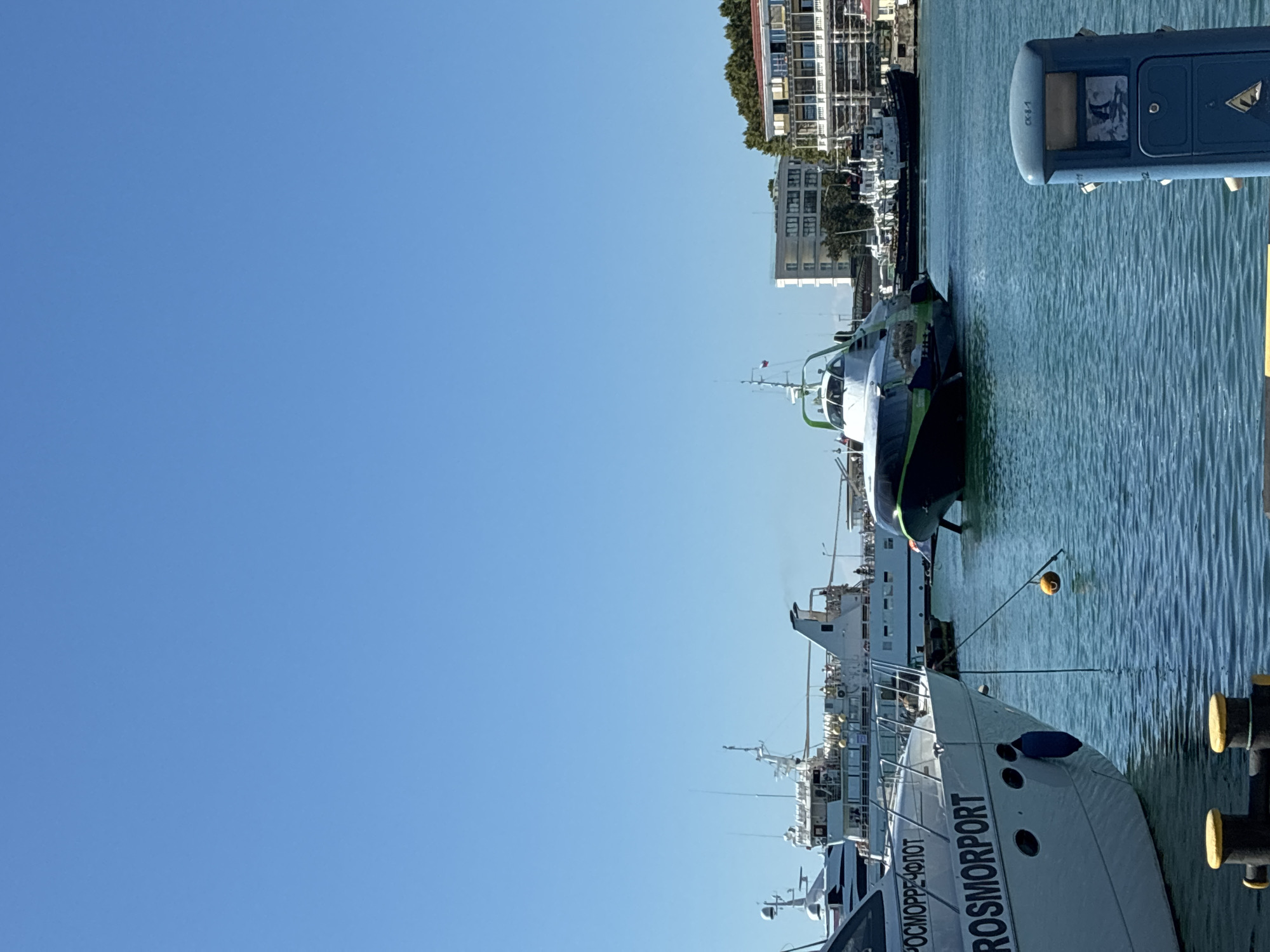 The image depicts a sunny day at a harbor in Sochi, Russia. In the foreground, a white yacht named "ROSMORPORT" is docked. To the right, a blue public utility bin is visible. In the middle ground, a green and white hydrofoil boat is underway, creating a wake. Several other boats and yachts are moored in the harbor. In the background, buildings and lush green trees line the shore. The sky is clear and blue.