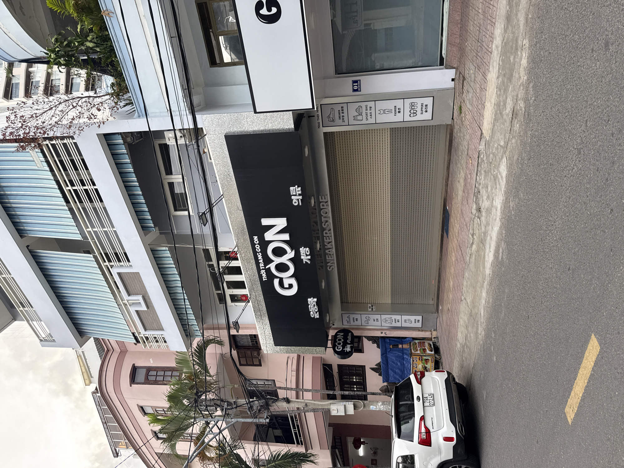 The image depicts a street scene in Nha Trang, Vietnam, featuring a row of multi-story buildings. The foreground shows a street with a white Kia Sorento SUV parked on the left side, displaying a license plate "79A 272245". Next to the SUV is a food cart with colorful food items displayed.
The buildings along the street present a variety of architectural styles and colors. The pink building on the left has several windows and balconies, with palm trees in front of it. Adjacent to it is a building with a gray facade, featuring a prominent black sign with white lettering that reads "GOON" in large font, along with Vietnamese and Korean text. Below this, "SNEAKER STORE" is visible. The storefront of this establishment appears to be a metal roller shutter. To the right of the "GOON" store, a white building with a black "G" logo is visible, and its entrance is a glass door. A vertical sign to the right of the "GOON" store lists various fashion items like sneakers, sport bags, fashion, and slippers, each with corresponding icons and text.
The buildings have balconies with awnings, and power lines are strung across the scene. The sky is overcast. The overall impression is of a commercial street in a Vietnamese city.
