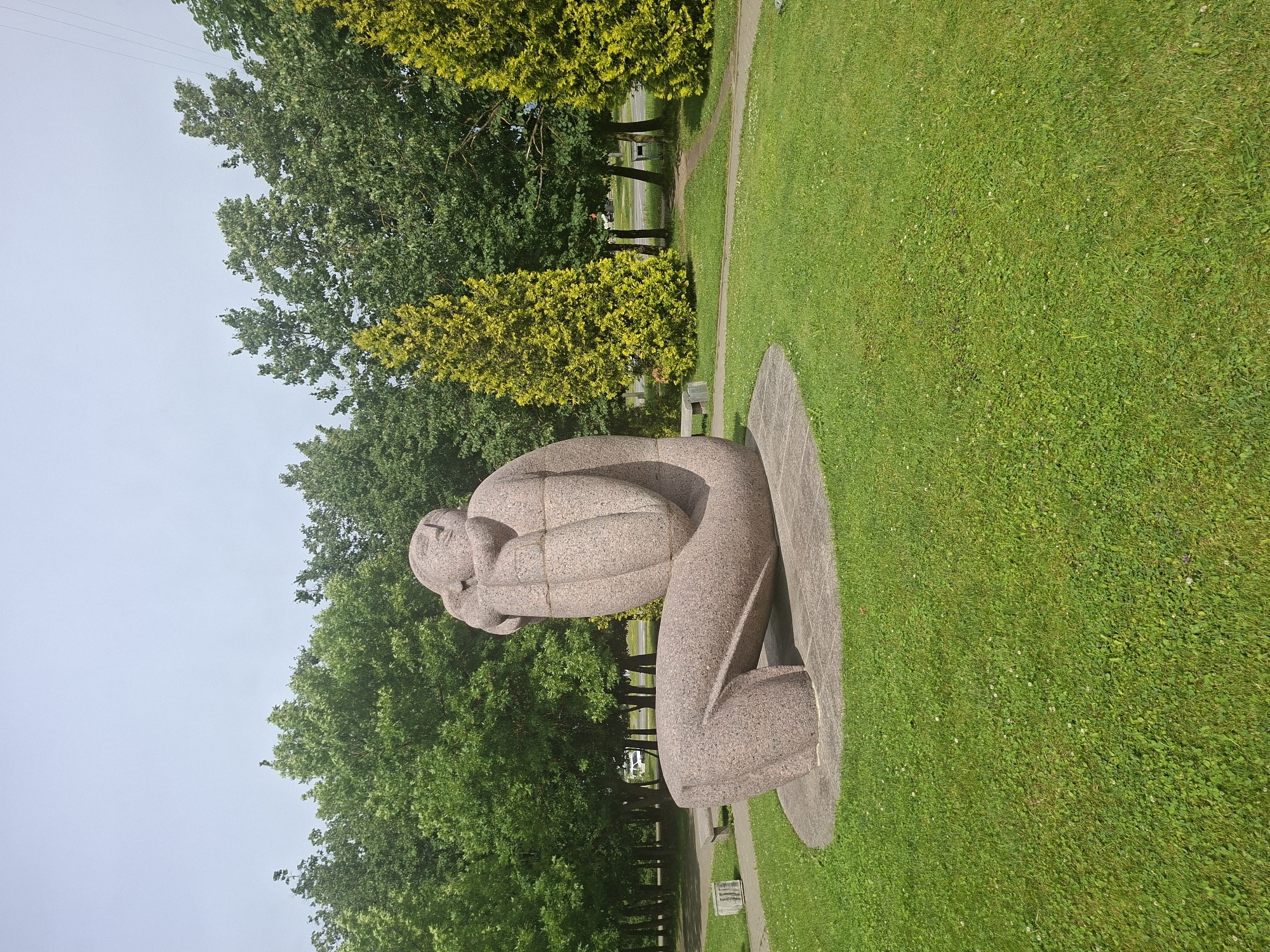 A stone sculpture of a person is set in a grassy park on a bright day. The sculpture is a stylized depiction of a figure curled into a fetal position, with its arms wrapped around its head. The stone is pinkish-grey and appears to be granite. The sculpture is situated on a concrete base, which is circular in shape.
The park has lush green grass, dotted with small white flowers and clover. Several trees with dense green foliage surround the sculpture, some with bright yellow leaves. A paved path runs through the park, bordered by more grass. In the background, a road can be seen with a few cars and a white structure that might be a building or a monument. The sky is overcast and light grey, suggesting a cloudy or misty day.
The overall scene conveys a sense of quiet contemplation or introspection, evoked by the posture of the sculpture and the peaceful park setting. The image appears to be taken in Kaunas, Lithuania. There is no visible text in the image.