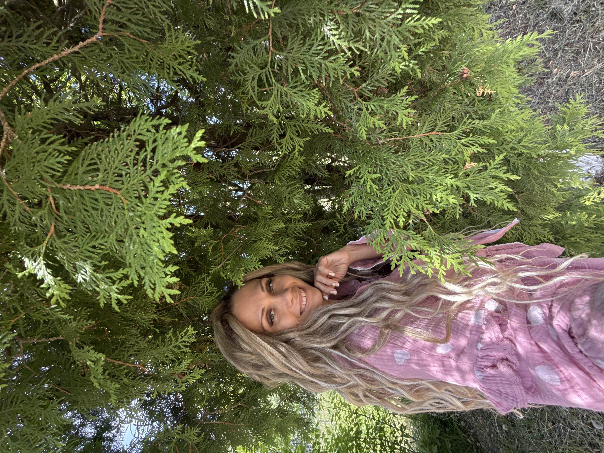 The image features a medium outdoor portrait of a woman with long, wavy, multi-toned blonde hair, including lighter, possibly braided or dreadlock-style strands. She has dark eyes, wears subtle eye makeup, and displays a broad smile, looking directly at the viewer. Her right hand, with manicured nails and light polish, is positioned with fingertips touching her chin. She is dressed in a light pink, textured garment, possibly made of crinkled cotton or gauze, adorned with irregularly spaced white heart patterns and horizontal ruffles across the chest area. The woman is positioned slightly to the left of the frame, partially enveloped by dense green foliage consistent with an evergreen shrub or tree, such as an Arborvitae (Thuja). Sunlight filters through the leaves, creating dappled lighting. In the background, additional green foliage and a small patch of blue sky are visible through the gaps in the leaves. The lower right foreground shows dry grass and bare earth. The image contains no visual elements to confirm a specific city or country.
