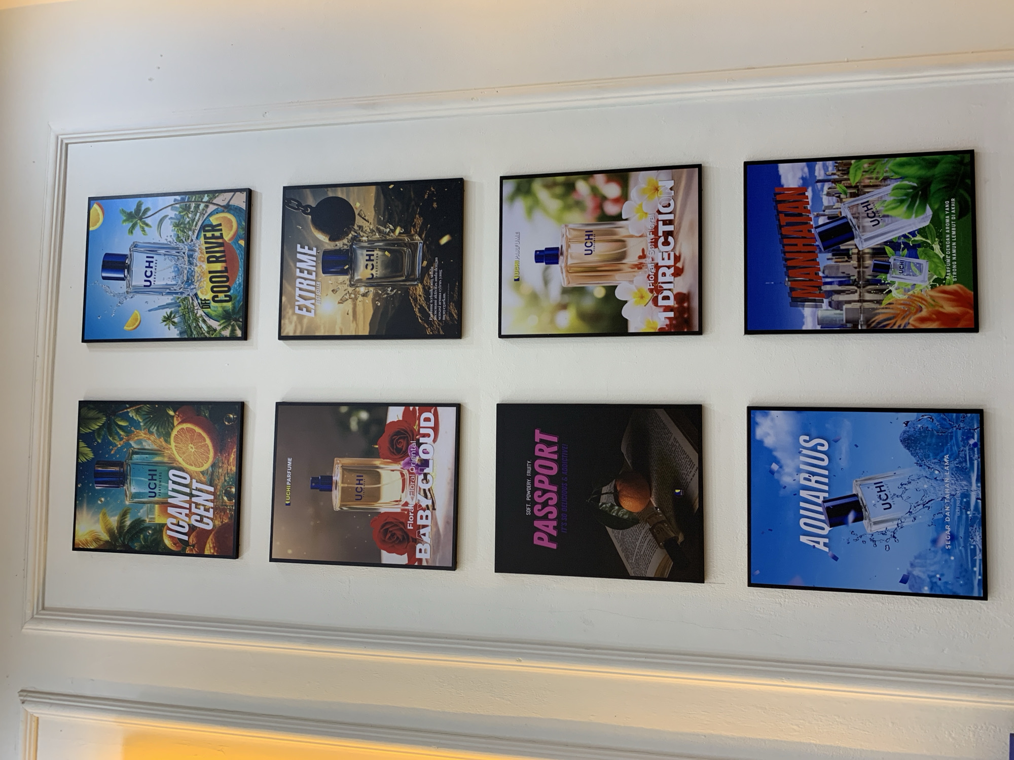 The image shows a white wall with several framed advertisements for "UCHI PERFUME" in Padangjaya, Indonesia. There are six advertisements displayed in a 3x2 grid. The top row features "ICENTO CENT" on the left and "COOL RIVER" on the right. The middle row displays "BABY CLOUD" on the left and "EXTREME" on the right. The bottom row shows "PASSPORT" on the left and "1 DIRECTION" on the right. In the lower portion of the image, two more advertisements are visible: "AQUARIUS" on the left and "MANHATTAN" on the right, also for UCHI PERFUME. The advertisements showcase perfume bottles with various backgrounds and descriptive text.