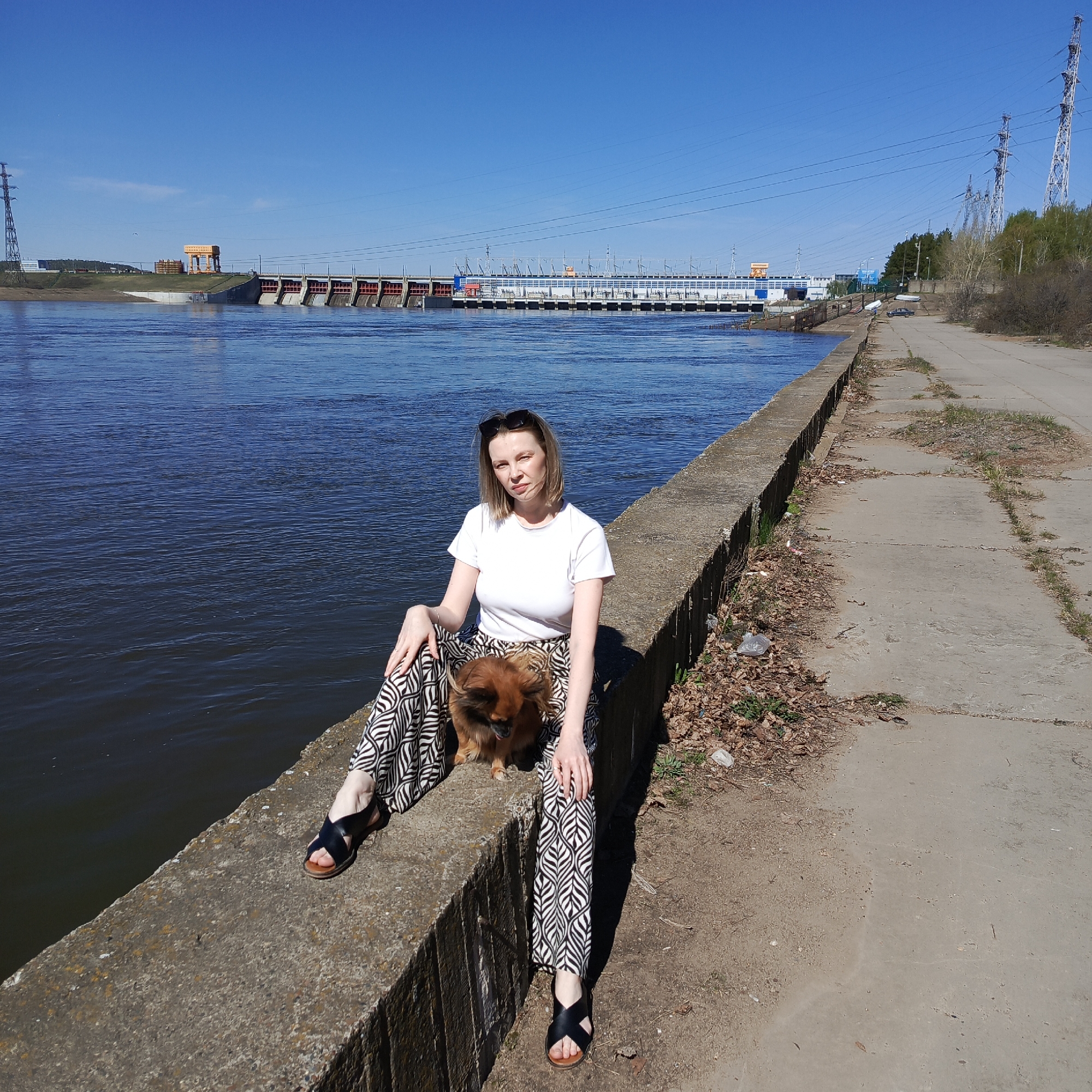 A woman and a small dog are seated on a concrete embankment in the city of Chaikovsky, Russia. The woman, with short blonde hair and wearing sunglasses, is dressed in a white t-shirt and patterned pants. She has dark sandals on her feet. A small, brown dog sits on her lap. Behind them is a wide river, reflecting the blue sky. In the background, a dam structure with various machinery and towers is visible. To the right of the embankment is a concrete walkway with weeds and debris along the edge. Power lines extend across the sky. The day appears to be sunny and clear.