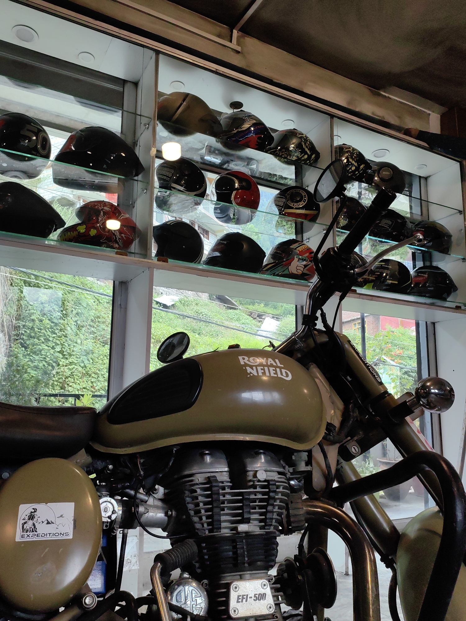 The image shows a Royal Enfield motorcycle parked indoors, likely in a showroom or workshop, in Vashisht, India. The motorcycle is a matte olive green color and features a "ROYAL ENFIELD" logo on the fuel tank. A sticker that reads "EXPEDITIONS" is visible on the side. In the background, a glass display cabinet showcases a variety of motorcycle helmets, some with intricate designs. The cabinet is illuminated, and the helmets are arranged on multiple shelves. Through a window behind the motorcycle, lush green foliage and a glimpse of buildings suggest an outdoor environment, typical of Vashisht's hilly terrain. The lighting is a mix of natural light from the window and interior lighting from the display case. There are no people visible in the image. The scene captures a collection of motorcycle-related items within a commercial setting in Vashisht.