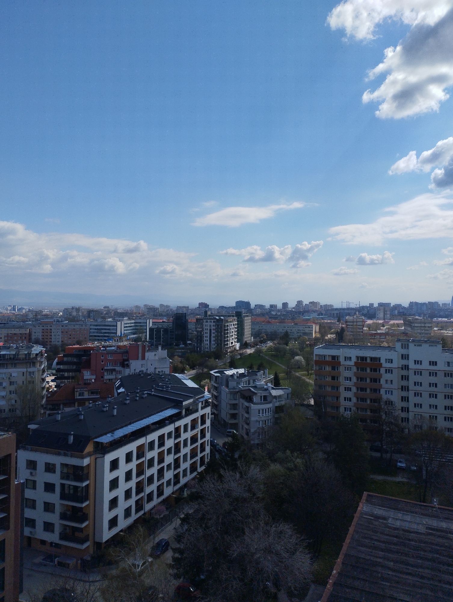 The image shows an aerial view of a city with a blue sky and scattered clouds. In the foreground, there are several buildings of varying heights and architectural styles, including modern apartment complexes with balconies and older structures. A small park with trees and green space is visible between the buildings. In the background, the city extends into the distance, with more buildings, possible mountains on the horizon. The location is Unknown, Unknown.