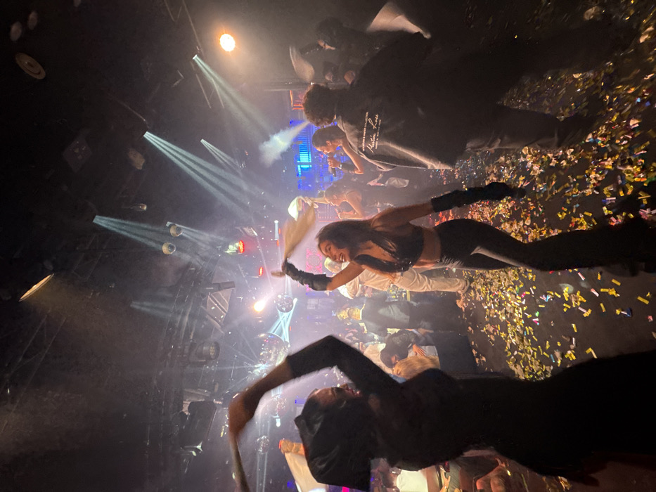 A vibrant nightclub scene is captured with spotlight beams cutting through a smoky atmosphere. A woman in a sequined crop top and pants, wearing gloves, smiles and waves a white fabric in one hand. Confetti showers the scene. Other figures are visible in the crowd, with individuals near the back appearing to perform or sing near illuminated screens. The perspective is tilted, adding to the chaotic energy of the event.