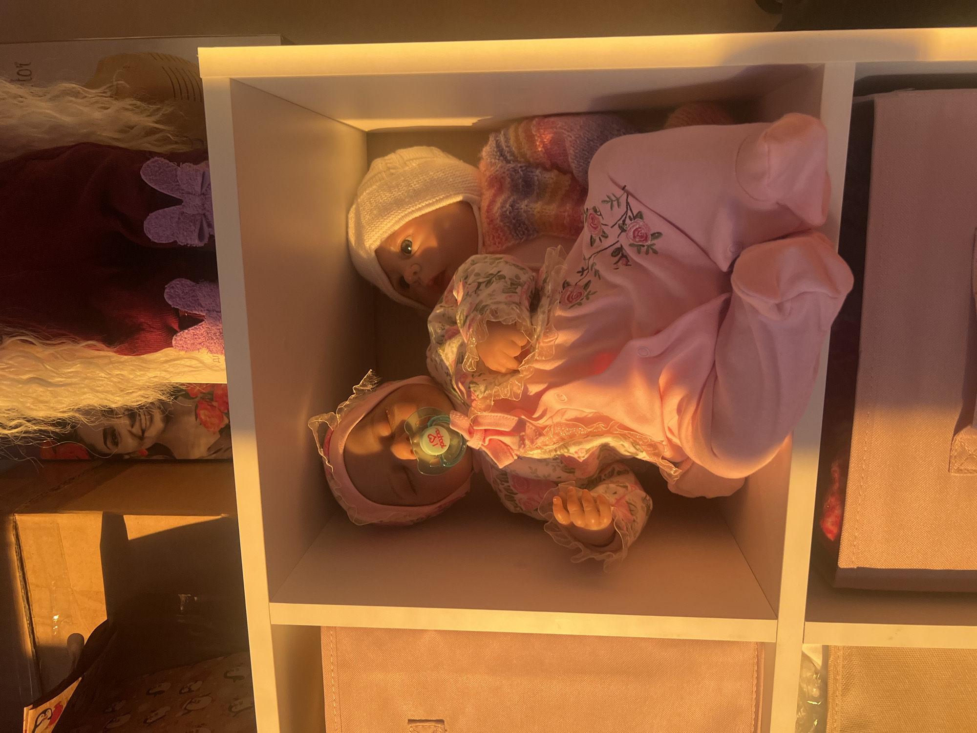 Two dolls are placed in a white shelf unit. One doll, wearing a white knit hat and a colorful sweater, is in a pink outfit with flower embroidery. The other doll has a pink bonnet and a pacifier and is wearing a floral-patterned dress with lace. The surrounding shelves contain boxes and a book or box with a face on it.
It is not possible to verify or determine the location in the city of Votkinsk, Russia from the given image.