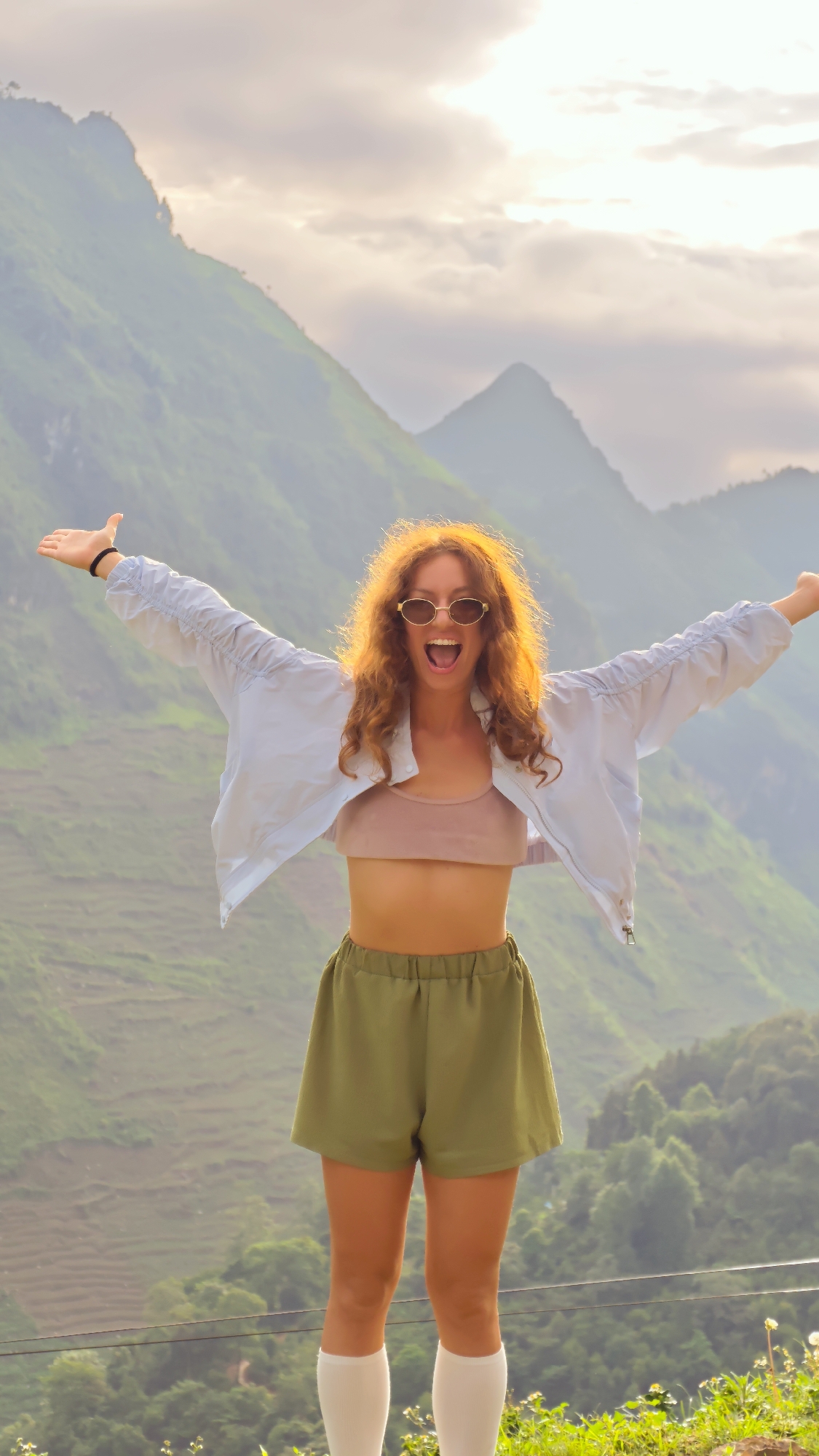 A young woman stands in a landscape with hills and mountains in the background. The woman is wearing a pink tube top, a light blue cropped jacket, and olive green shorts. She also has white knee-high socks and round sunglasses. She is smiling with her mouth open and her arms outstretched. The setting is likely Lũng Cú Commune, Vietnam, given the mountainous backdrop and the lush vegetation. The sky is overcast.