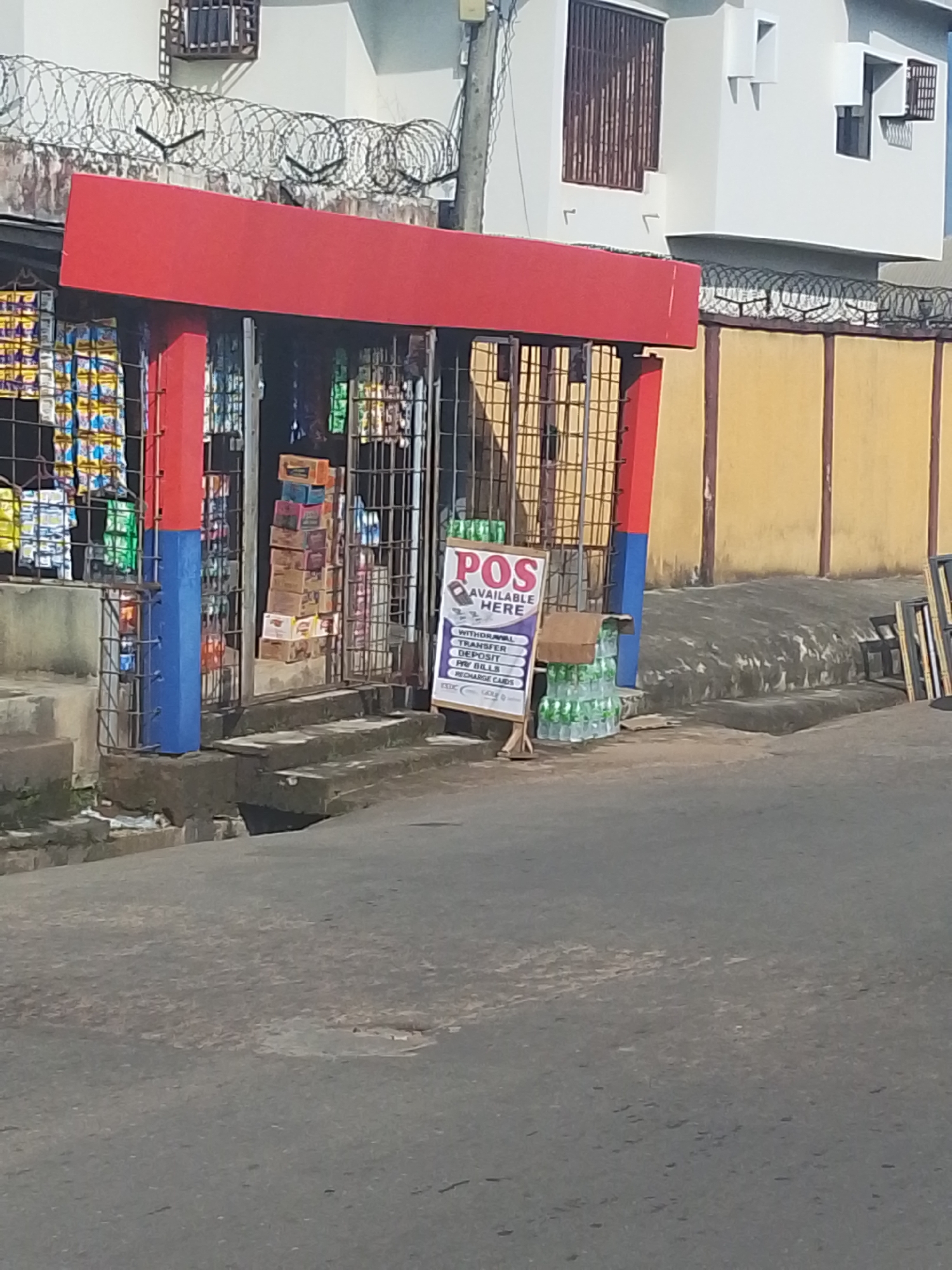 The image displays a street view in Umuahia, Nigeria, during daylight hours, focusing on a small retail shop and its surroundings. The shop has a distinctive red and blue painted facade, protected by metal grates, and features concrete steps leading up to its entrance from the asphalt road. Inside, various packaged goods, including snacks and drinks, are visible on shelves and stacked.
A white sign on a wooden stand outside the shop prominently advertises "POS AVAILABLE HERE." Below this, a list of financial services is displayed, including "WITHDRAWAL," "TRANSFER," "DEPOSIT," "PAY BILLS," "RECHARGE CARDS," and "CABLE TV." An "OPAY" logo is also visible at the bottom of the sign. Several packs of bottled water are stacked on the ground beside the sign.
Behind the shop, a yellow-painted wall is topped with coils of barbed wire. Further back, a modern white building with brown slatted window coverings and more barbed wire on its perimeter is visible. The bright lighting suggests a sunny day. The asphalt road in the foreground shows signs of wear. On the right, near the yellow wall, stacked metal frames or objects are present. No people are visible in the scene.