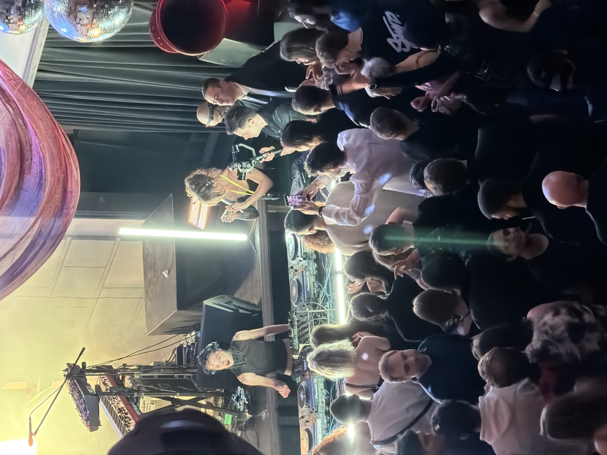 The image depicts a performance in a crowded venue, likely a nightclub, in Tbilisi, Georgia. A female DJ in a black top and headphones is performing on a stage with Pioneer DJ equipment. To her left, a woman plays a keyboard, illuminated by stage lights. The audience is densely packed, with many people filming or photographing the performance on their phones. Above, there are disco balls and red cylindrical objects hanging from the ceiling, which is draped with black fabric. A large, colorful fabric installation hangs on the left side of the frame. The overall lighting is a mix of stage lights and dim ambient lighting.