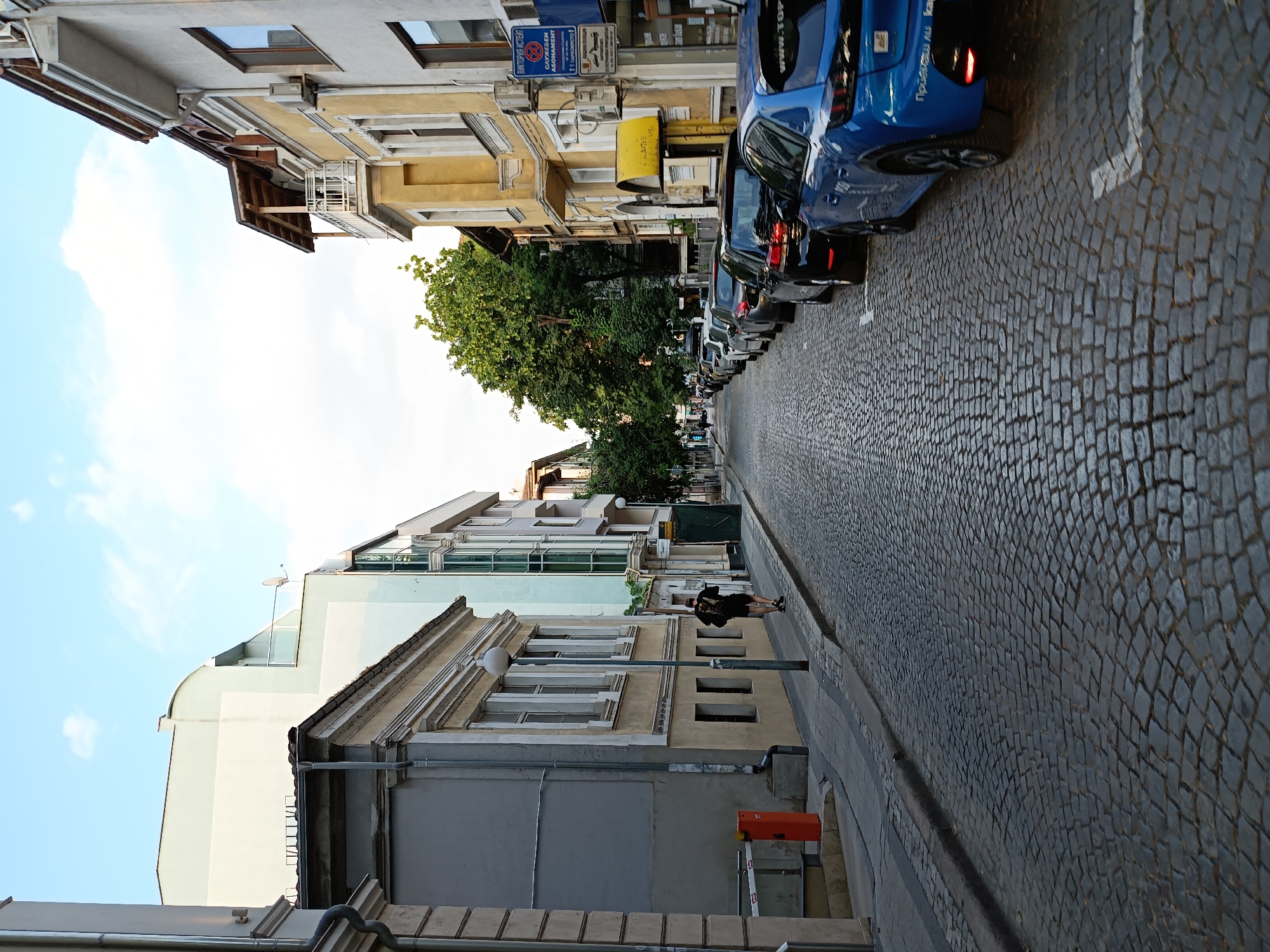 The image depicts a street scene in Plovdiv, Bulgaria, with buildings lining one side of a cobblestone road. The perspective is from street level, looking down the street towards a row of parked cars. The buildings vary in architectural style and color, with some featuring ornate detailing and balconies, while others appear more modern. Trees with lush green foliage are visible between some of the buildings.
A person is standing on the sidewalk, facing away from the camera and looking at their phone. The time of day appears to be late afternoon or early evening, suggested by the soft lighting and shadows. The sky is visible in the upper left quadrant of the image and is a light blue with some wispy clouds.
Several cars are parked along the curb. The closest car is a blue hatchback. The road surface is made of dark, uneven cobblestones, with white parking lines painted on it. The overall atmosphere of the scene is calm and ordinary, typical of a residential or mixed-use street in a European city. Some visible text, likely signs or labels on buildings or cars, is present but illegible due to the resolution and angle.
