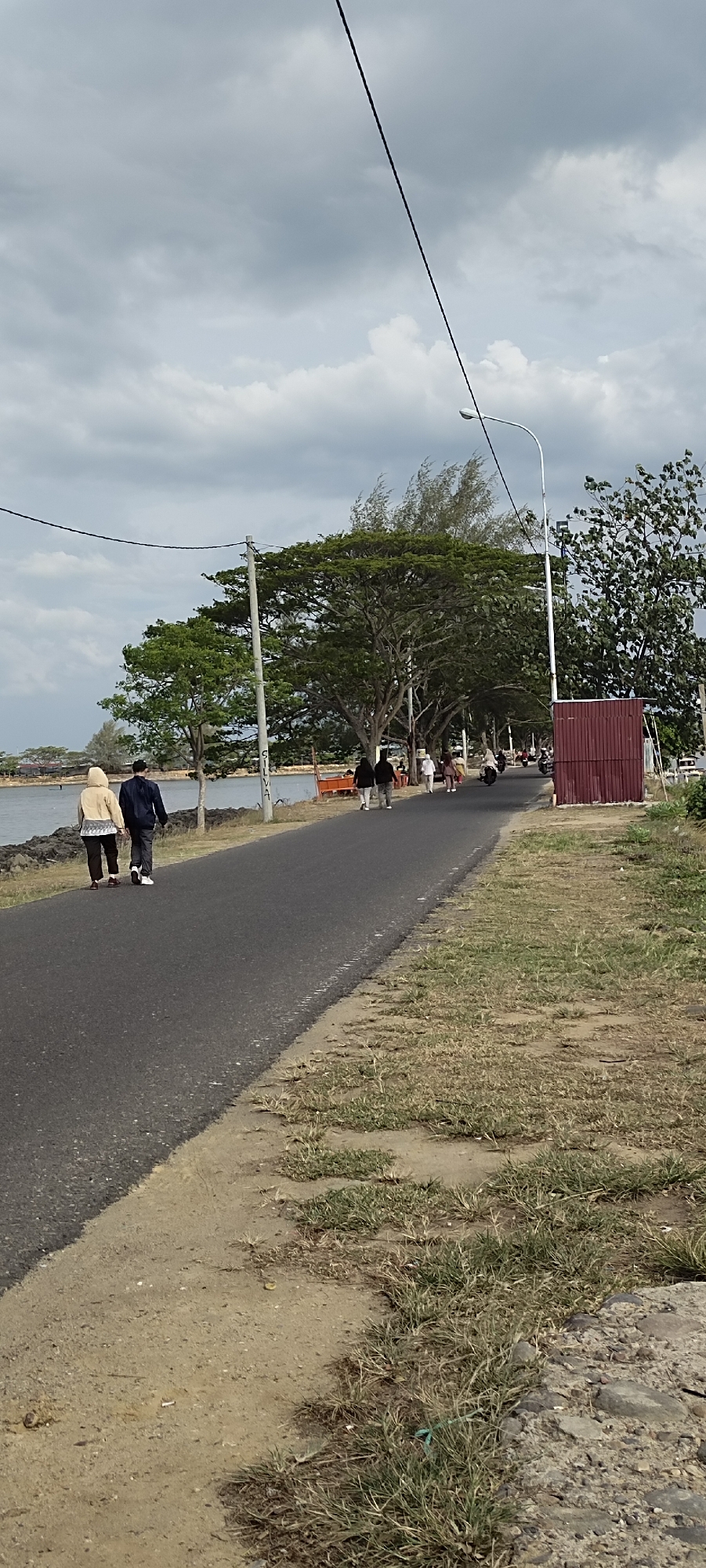 In Lhokseumawe, Indonesia, a paved road runs alongside a body of water, possibly a river or bay. The road is bordered on one side by the water and on the other by a grassy embankment and trees. The sky is overcast, with dramatic clouds. Several people are walking or riding motorcycles along the road. In the foreground, two individuals are walking away from the camera, one in a light-colored hooded jacket and the other in a dark jacket. Further down the road, other pedestrians and motorcycles are visible. A small, red, corrugated metal structure stands on the right side of the road. Power lines crisscross the sky above the scene.