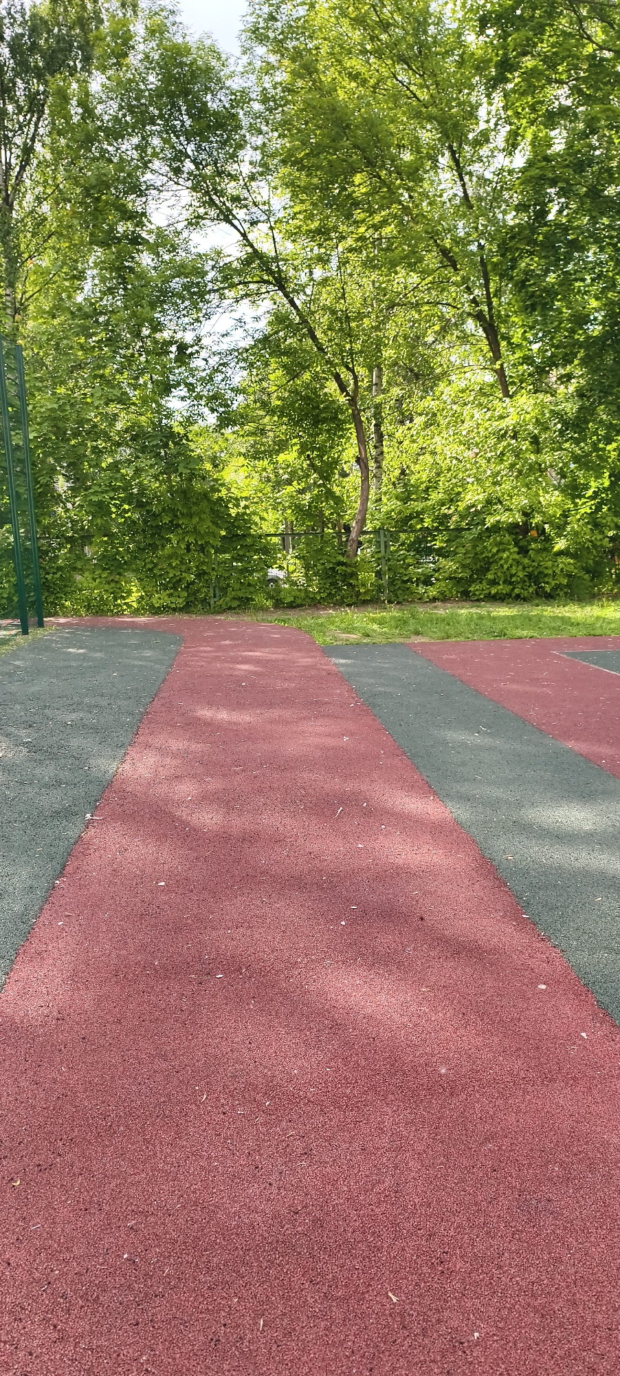 The image captures an outdoor recreational area on a bright, sunny day. The foreground features a rubberized surface, primarily red with a long grey stripe running down the center towards the background. Another grey section is visible on the left side, curving slightly. Small white specks, possibly fallen leaves or debris, are scattered across the red surface, and subtle shadows suggest overhead foliage.
In the midground, beyond the patterned surface, there is a low metal fence or railing, largely obscured by lush green bushes and dense foliage. To the far left, a green metal pole, possibly part of a pull-up bar or other playground equipment, is partially visible.
The background is dominated by numerous tall trees with vibrant green leaves, indicating a summer or late spring setting. Glimpses of a bright, clear sky can be seen through the tree canopy. The overall scene is peaceful and devoid of people or specific activities, suggesting a quiet moment in a park or playground setting. No visible text is present.