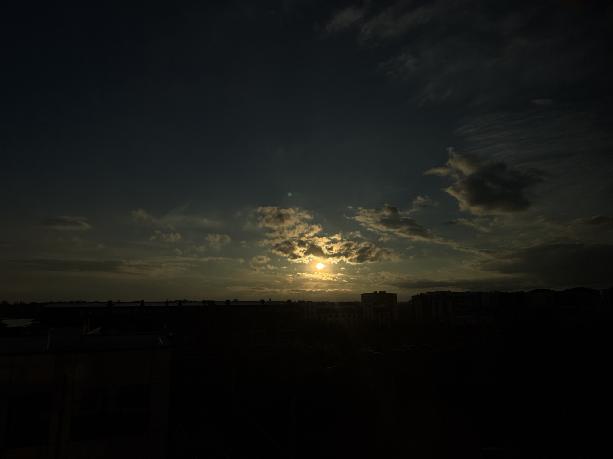 The image depicts a city skyline at dusk, with the sun setting behind clouds. The sky is a deep blue, transitioning to lighter shades of yellow and orange near the horizon. The sun itself is a bright, circular orb, partially obscured by wispy clouds. A line of buildings, silhouetted against the sky, forms the urban landscape. The foreground is dark, with hints of structures and possibly vegetation. The overall impression is one of a peaceful, atmospheric evening in an urban setting.