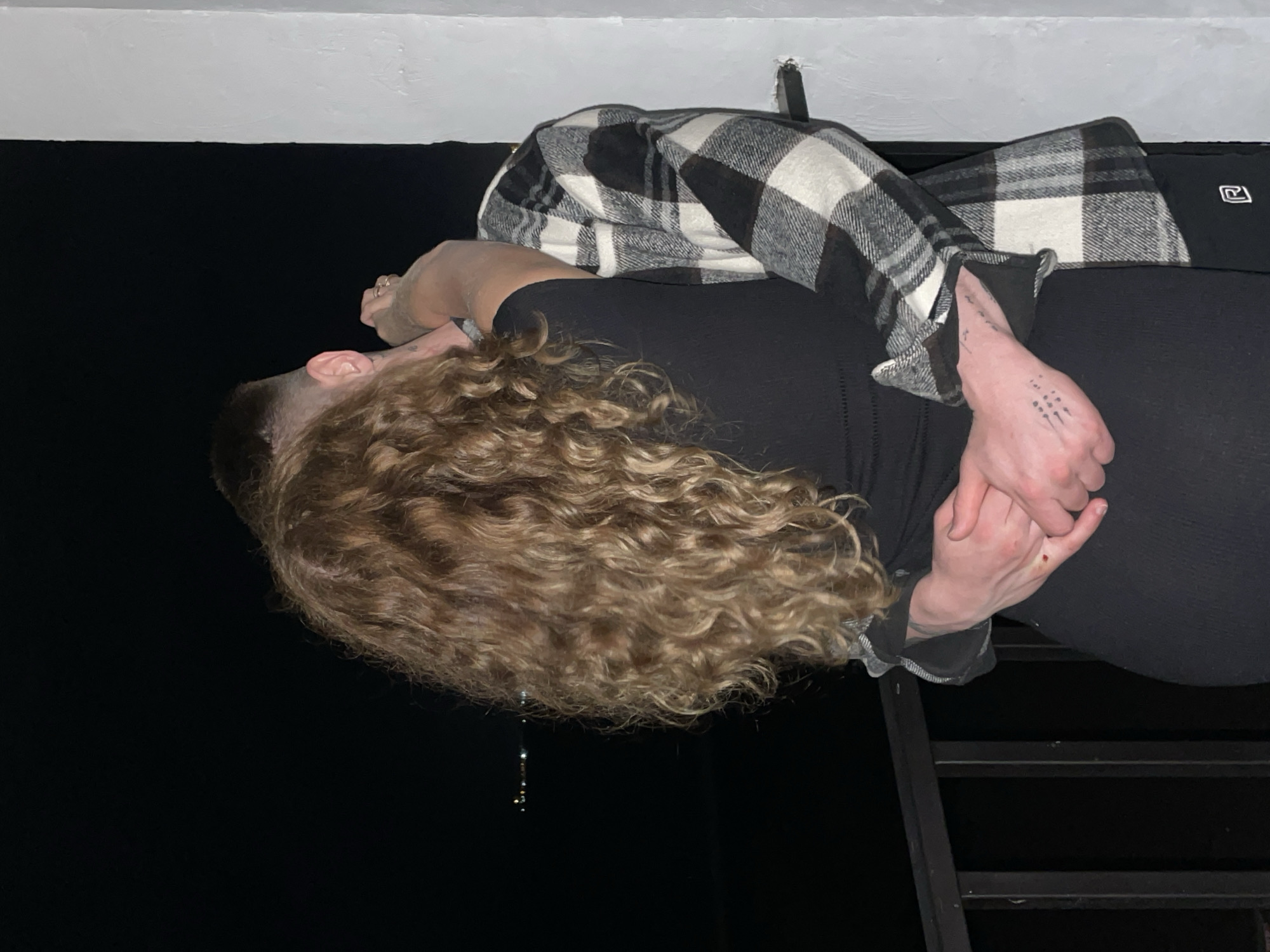 A person with long, curly blond hair is positioned near a metal ladder in an environment with dark background. The person is wearing a black top and black bottoms with a black and white plaid shirt. Their hands are clasped together, and there are tattoos visible on the hand. The location appears to be indoors, as a white ceiling is visible.
