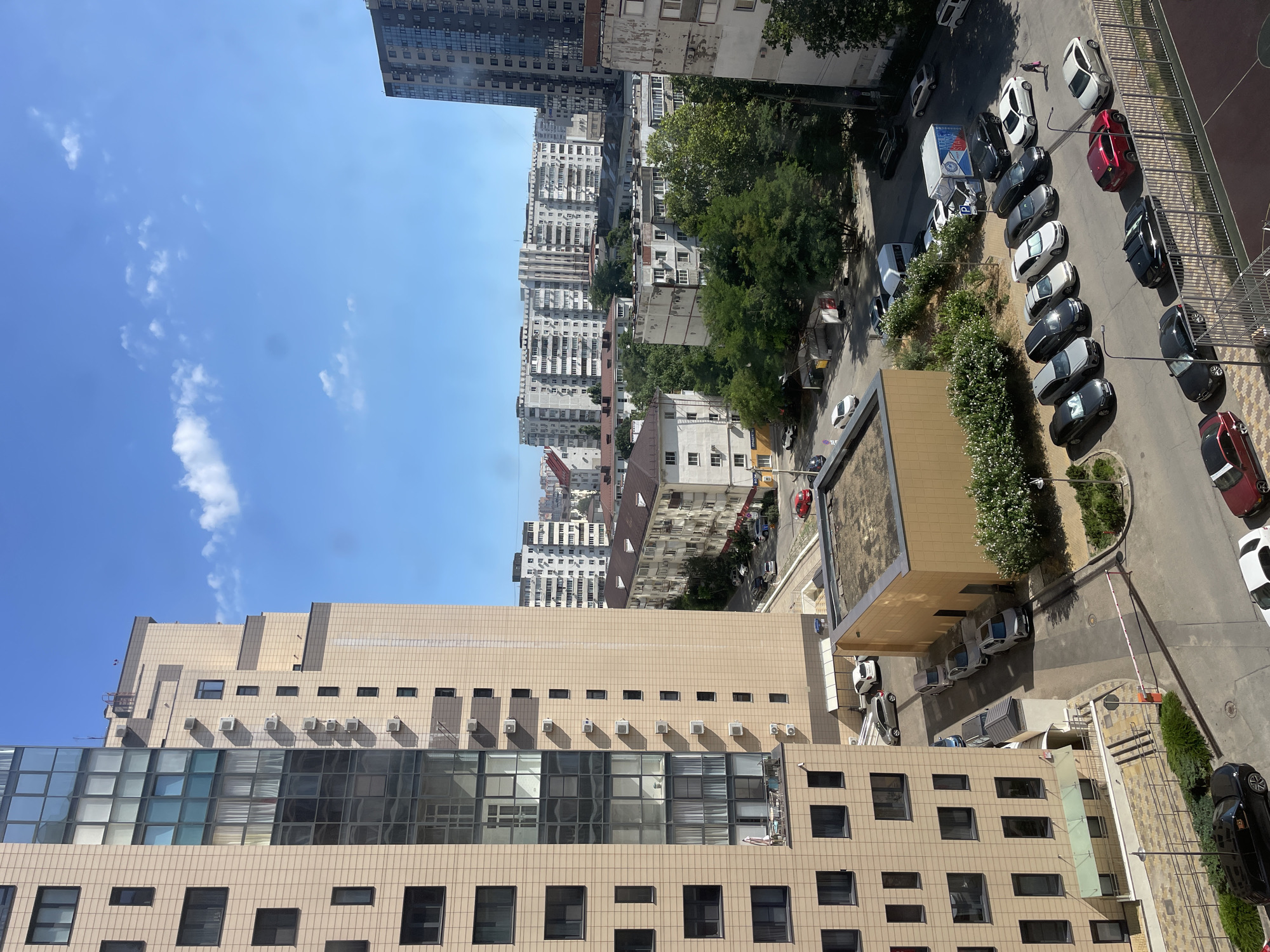 This is an aerial, eye-level shot of a city block in Novorossiysk, Russia, on a bright, sunny day with scattered clouds. In the foreground, a modern, tan-colored apartment building dominates the left side of the frame. Its facade features a grid of dark windows, and balconies with outdoor furniture are visible on a lower floor. Adjacent to this building is a smaller, yellow-clad structure.
The scene depicts a parking lot filled with numerous cars of various colors and models, parked in a row. Among them are a black sedan, a silver SUV, several white cars, a red coupe, and a dark gray sedan. A person in dark clothing is walking across the parking lot. A white van with blue and red lettering is parked in the lot, and other vehicles, including a white minivan and a white van with a blue box body, are also present.
Behind the parking lot, several other residential buildings are visible. Some are older, grey concrete structures with numerous windows, while others are more modern, with balconies and darker facades. Lush green trees are scattered throughout the cityscape, interspersed between the buildings and lining some of the streets. The sky is a clear blue, with a few wispy white clouds. The overall impression is of a dense urban environment with a mix of modern and older architecture, and signs of everyday life with parked cars and pedestrian activity.