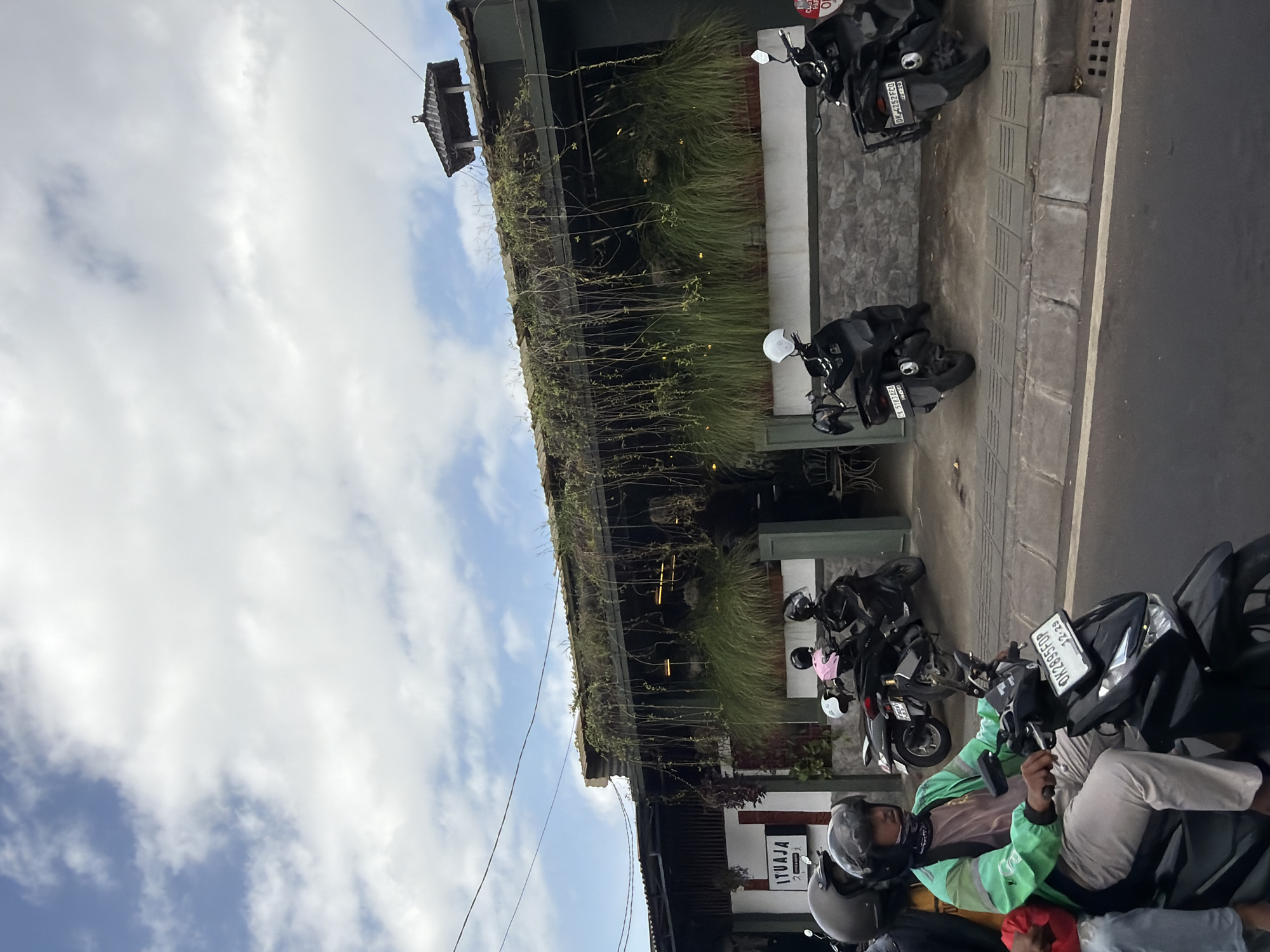 The image depicts a street scene in Kuta Selatan, Indonesia, with a focus on a building facade adorned with hanging plants and a prominent sign that reads "ITUJAJA". Several motorcycles are parked along the sidewalk in front of the building. In the foreground, a person wearing a green jacket and a helmet is riding a black motorcycle, with another individual partially visible behind them. The sky above is partly cloudy with blue patches. The building appears to be a commercial establishment, possibly a cafe or restaurant, given the outdoor seating visible through the windows. The overall environment suggests a bustling urban setting, common in tourist areas like Kuta Selatan.