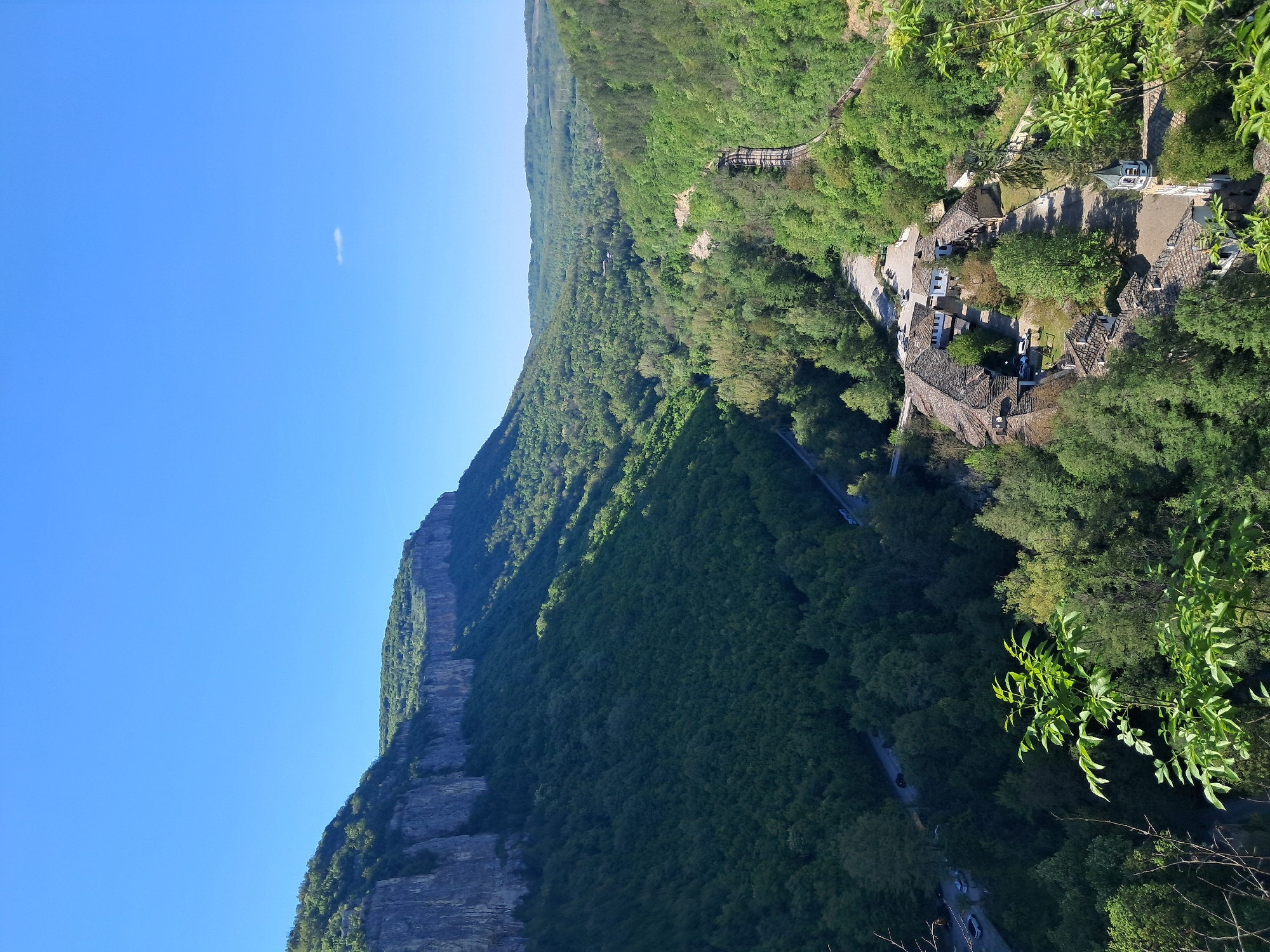 A high-angle shot depicts a mountainous landscape, likely in the vicinity of Gesha, Bulgaria. A sheer cliff face dominates the left side of the frame, transitioning into a dense forest covering the lower slopes. A winding road is visible through the trees. On the right side, a collection of buildings with tile roofs suggests a small settlement or village. A few cars are parked near these buildings. The sky is clear and blue with a small puff of clouds, and the lighting indicates a sunny day. The overall scene presents a tranquil rural environment with a combination of natural and man-made elements.