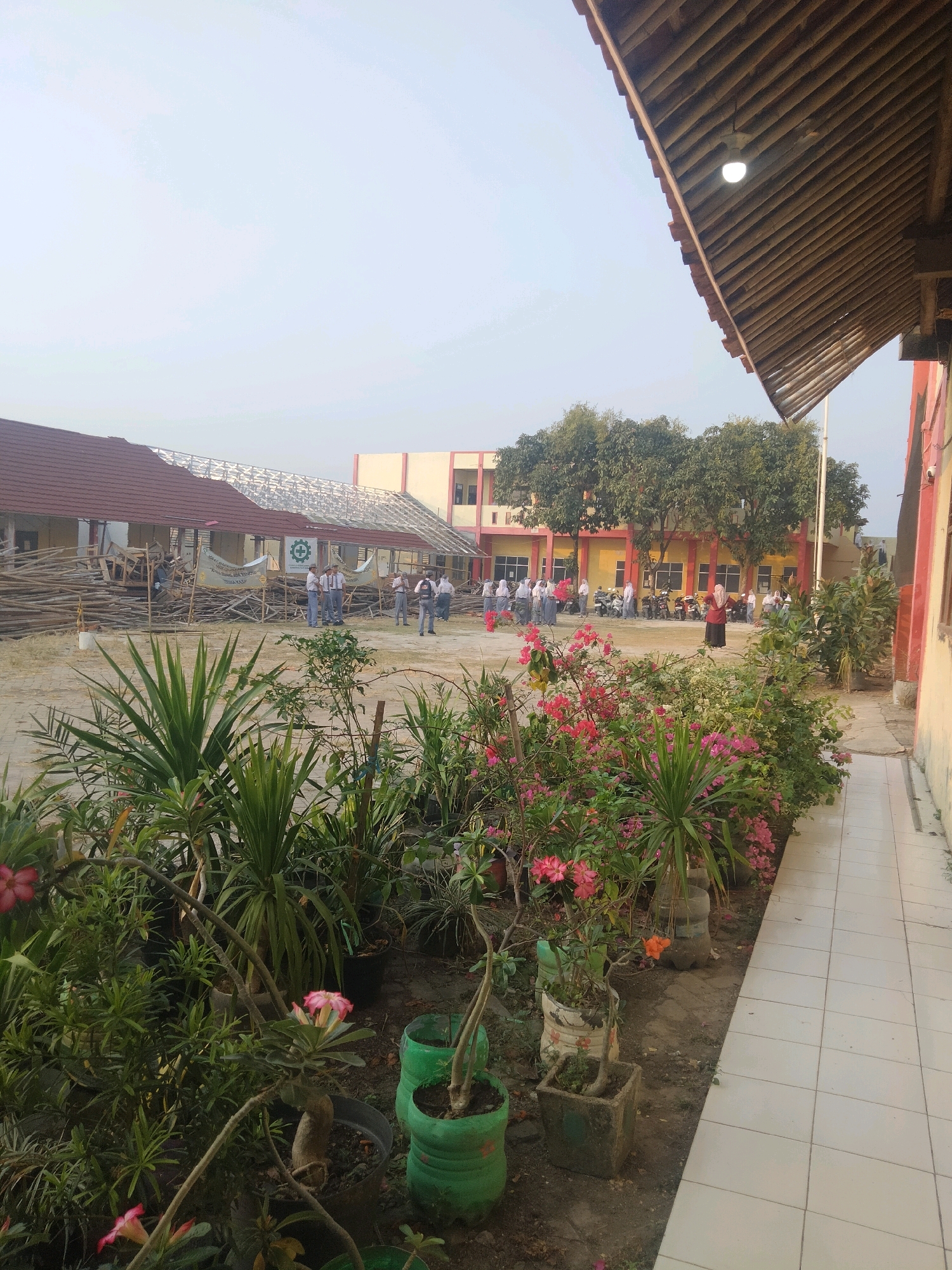 In Suka Sari, Indonesia, a schoolyard is captured in a wide-angle shot, focusing on the foreground's lush greenery and the mid-ground's school buildings. A paved walkway runs along the right side of the image, leading towards the foreground's dense foliage of flowering plants and tropical greenery, some of which are in repurposed plastic containers.
In the mid-ground, a school building under construction is visible, with scaffolding and piles of lumber. Several students in school uniforms are gathered in the vicinity, some appearing to observe the construction. Another school building, painted yellow and pink, stands in the background, with trees in front of it. A group of students and a few motorcycles are present in front of this building, suggesting a typical school day. The sky is clear and bright, indicating daytime.
