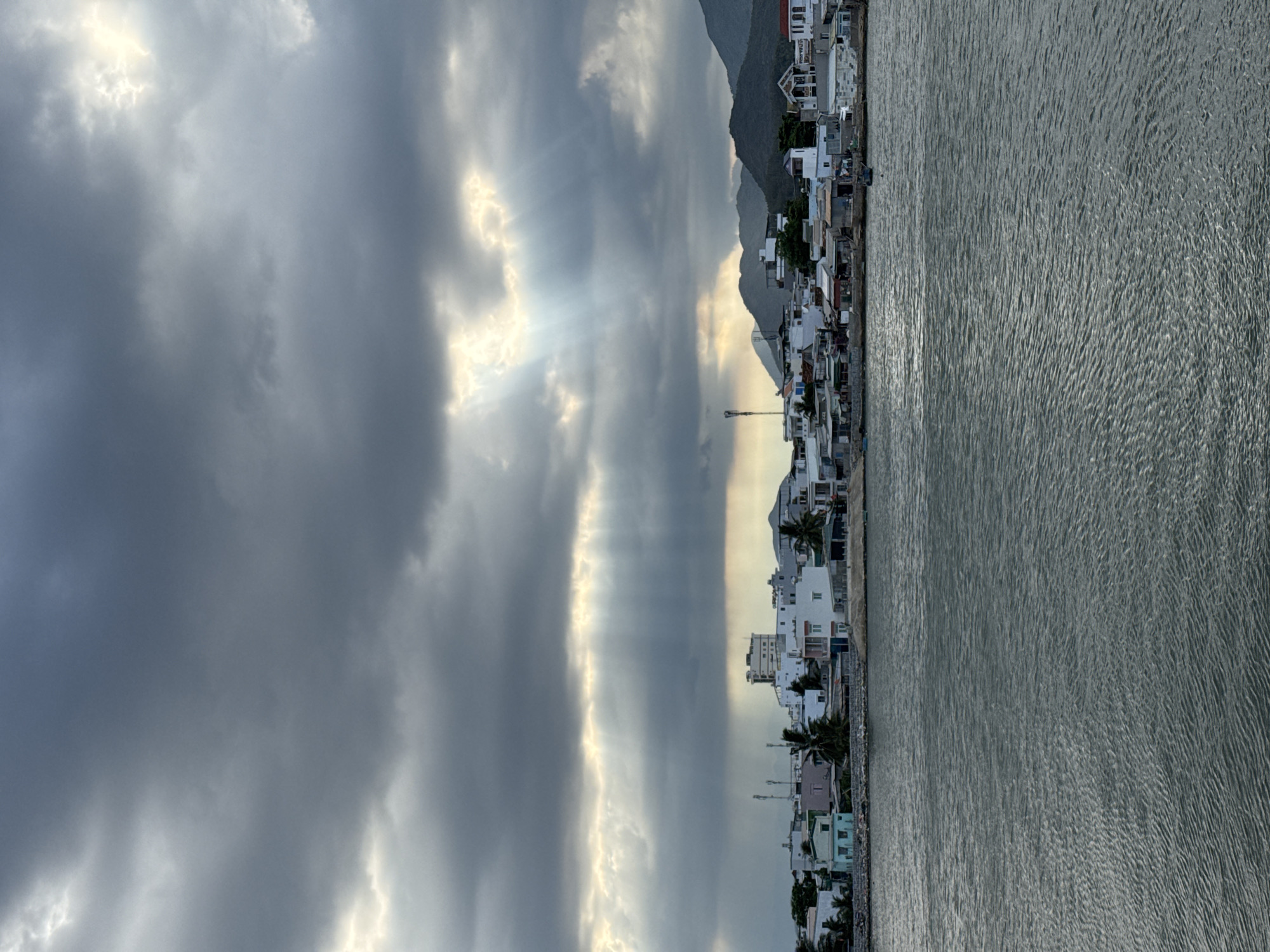 The image depicts a coastal scene in Nha Trang Ward, Vietnam. The right side of the image shows the water, with visible ripples and reflections. Adjacent to the water is a line of buildings, characterized by white and other pale colors, with varied architectural styles. A mountainous backdrop is visible behind the buildings, partially obscured by a lighter sky. The sky is filled with heavy, gray clouds, with streaks of light breaking through, creating a dramatic effect. The image is rotated.