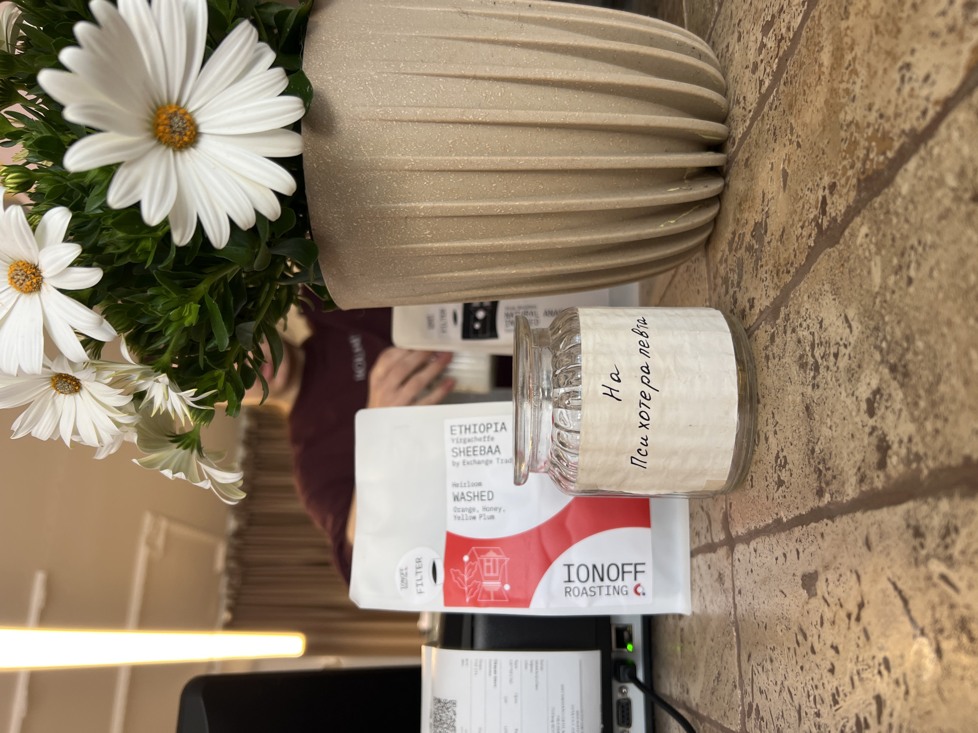 The image shows a table with a bouquet of white daisies in a beige vase. Next to the vase is a glass jar with a handwritten label in Cyrillic, "На Психотерапевта," which translates to "For the Psychotherapist." Below the jar is a white package of "IONOFF ROASTING" coffee from Ethiopia, specifically "SHEEBAA" Virgacheffe, described as "WASHED" with "Orange, Honey, Yellow Plum" flavors. Below the coffee package, a white receipt is visible. A black computer or monitor is also present. A person's arm wearing a maroon shirt is in the background. The table's surface appears to be textured and patterned. The scene is in Bar, Montenegro.