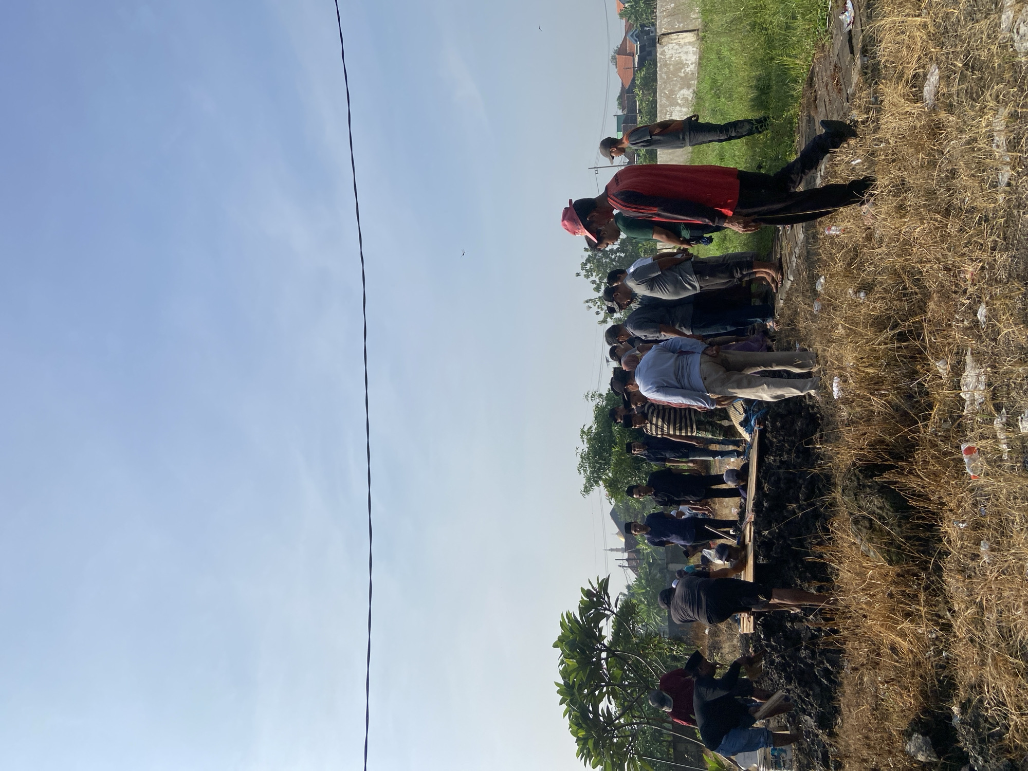 A group of men are gathered outside, likely participating in some kind of communal activity. They stand on a narrow patch of ground between a dirt bank covered with dry grass and scrub, and what appears to be an open, recently dug area. Some of the men are bending over as if in prayer or contemplation. The backdrop features a clear blue sky with a single visible power line. Further in the background, low buildings can be seen, suggesting a residential or suburban setting. The location is Kedondong, Indonesia.