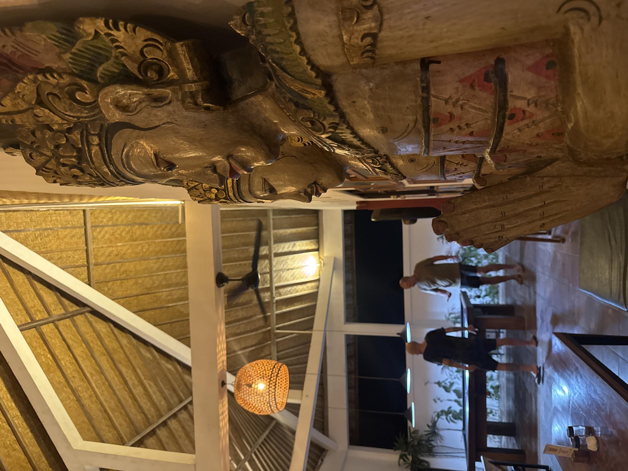The image shows an indoor dining area. In the foreground, a large, carved wooden sculpture of a figure with elaborate details dominates the view.
In the background, two individuals are standing near tables. One person is wearing a dark shirt and shorts, while the other is wearing a light-colored shirt and shorts. The dining area is lit by pendant lights, one of which is a woven basket-style lamp. The ceiling is high, with exposed beams and a fan. Large windows reveal a dark outdoor setting with some plants. The location is in the city of Unknown, Unknown.