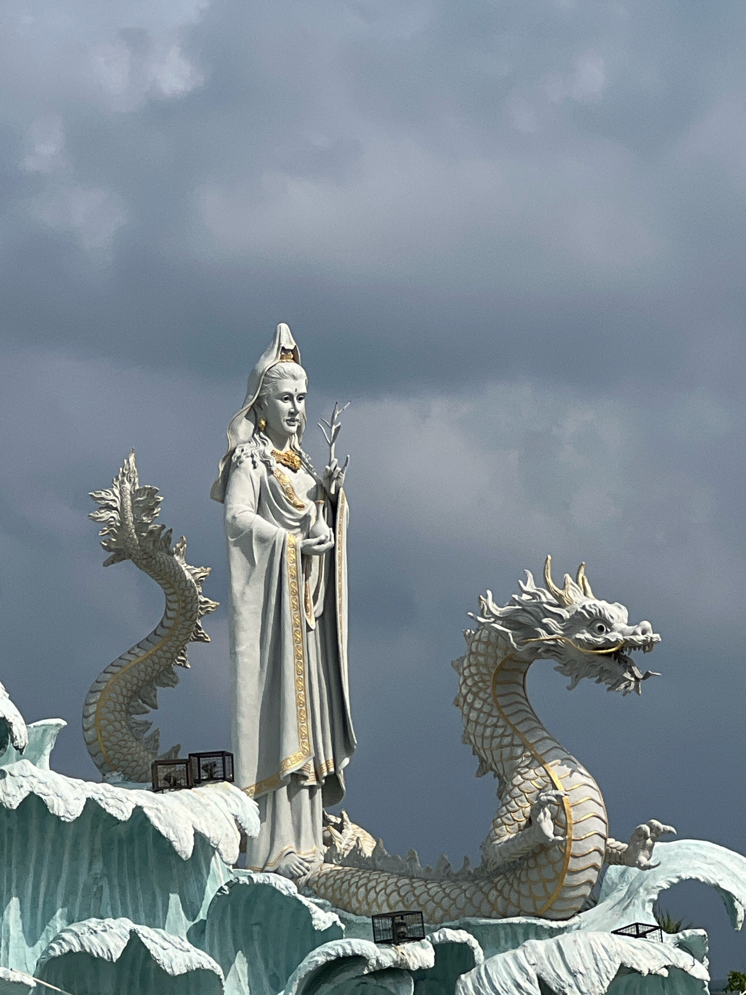 This image shows a large white statue of a woman in traditional Asian attire, standing between two white dragon statues. The woman is wearing a tall headdress and a flowing robe with gold trim. She holds a leafy branch in her right hand and what appears to be a small vase or vessel in her left. The dragons are detailed with scales and ornate golden accents. They are positioned as if emerging from stylized waves, which form the base of the sculpture. The background consists of a dark, cloudy sky. This sculpture is located at the Vihara Dharmayana Kuta in Kuta, Indonesia.