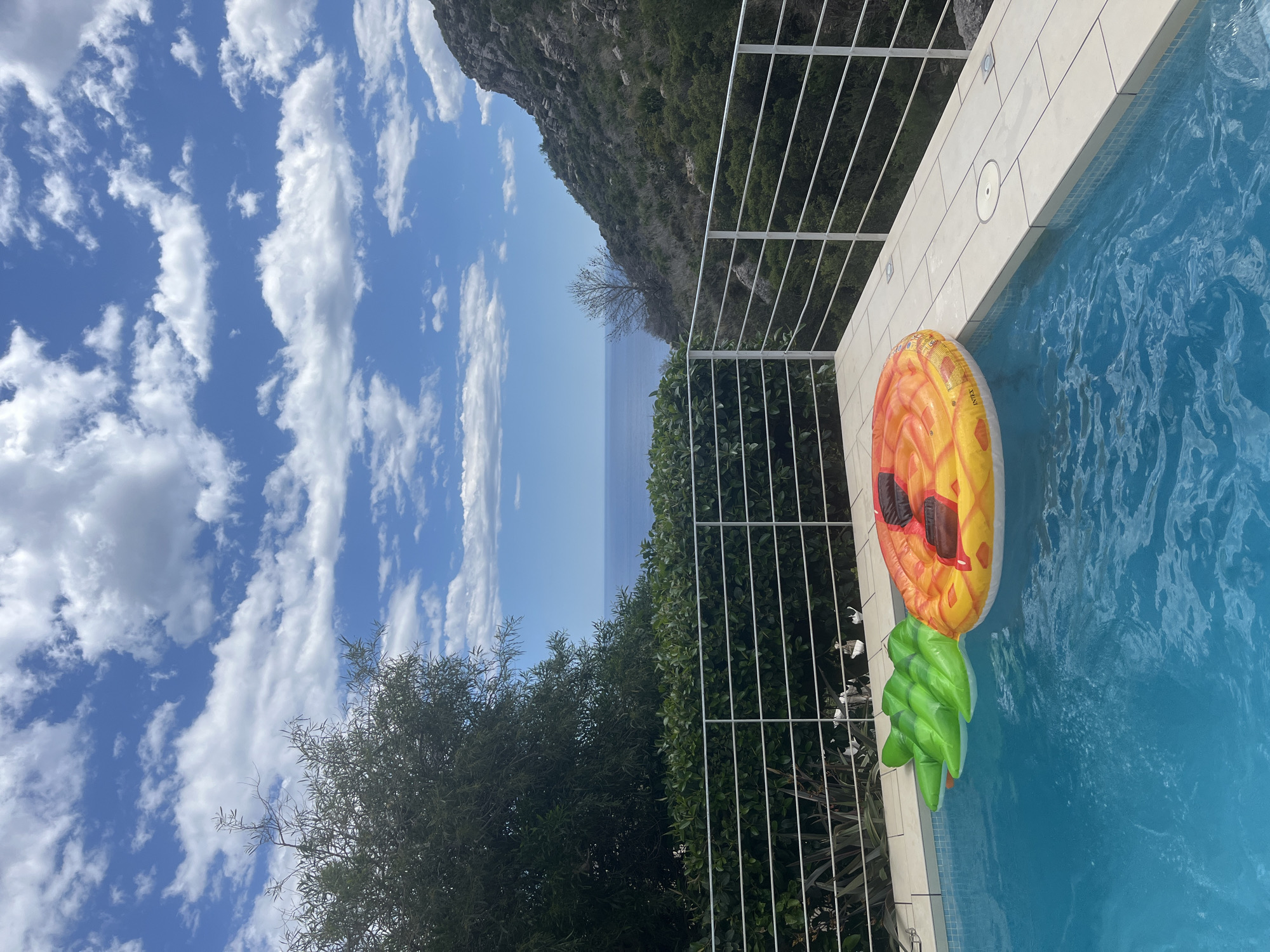A slightly rotated, medium shot depicts a section of a swimming pool with an inflatable pineapple float on a sunny day in el Poble Nou de Benitatxell / Benitachell, Spain. The pool is filled with clear blue water. The pineapple float has a yellow and orange body, green leafy top, and brown sunglasses. A white metal railing separates the pool from greenery and a cliff. Beyond the railing, one can see the ocean and the horizon, above which there is a bright blue sky filled with fluffy white clouds. A tree is visible in the foreground on the left, with dark green foliage. The setting sun casts shadows on the water and the pool deck.