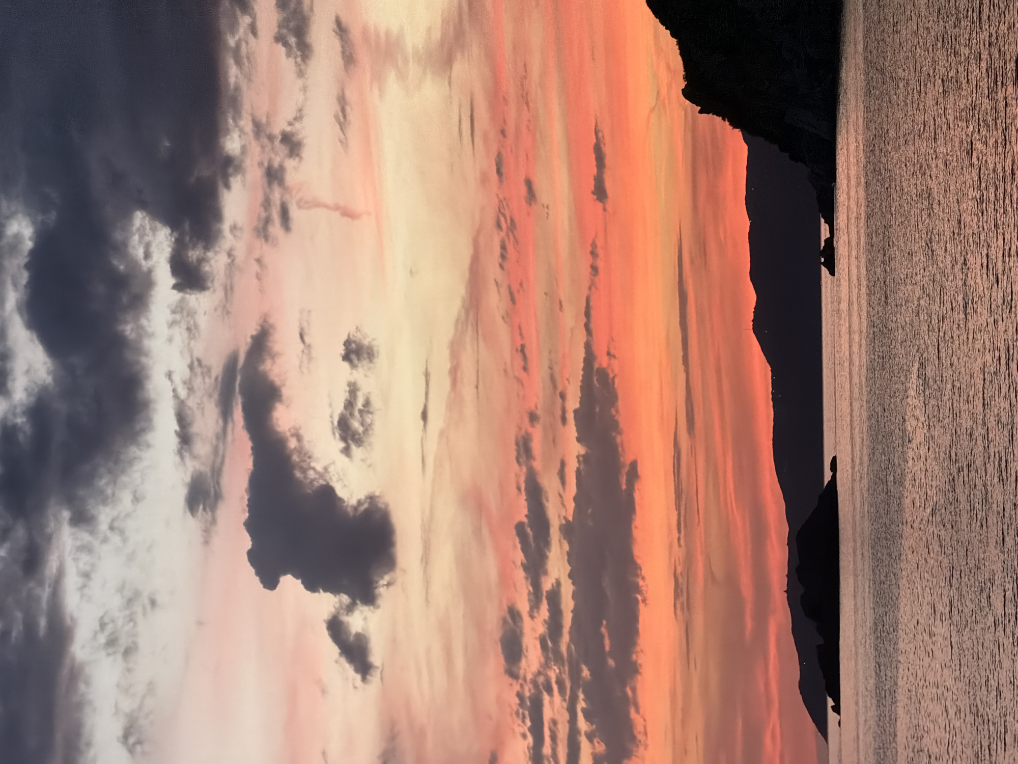 The image captures a twilight seascape in Perazića Do, Montenegro, characterized by a vivid, multi-hued sky and dark, silhouetted landforms against the water.
The sky, occupying the upper two-thirds of the frame, displays a pronounced color gradient. The uppermost portion is a light, muted gray transitioning into soft pink and pale yellow. Below this, the sky intensifies, featuring broad bands of vibrant orange and deep red that extend towards the horizon. Scattered throughout the sky are numerous clouds. In the upper half, dark gray, dense cloud formations are prominent, with one large, irregular cloud mass in the upper left-center area. Further down, more fragmented and elongated dark gray clouds are interspersed among the fiery sky colors. A thin, vertical, wispy streak, possibly a contrail or unique cloud formation, is visible in the mid-right sky.
The lower third of the image is composed of the sea and land silhouettes. In the mid-ground of the water, a prominent, dark, rocky island or a cluster of small islands is sharply silhouetted against the bright horizon. A tiny, indistinct structure, possibly a lighthouse or beacon, is discernible on the highest point of this landmass. Behind this foreground island, a continuous line of darker, lower hills or distant coastline stretches across the background, exhibiting subtle undulations and faint light points that suggest distant structures. On the immediate right, a steep, rugged, dark cliff face juts into the frame, extending vertically down to the water.
The surface of the sea reflects the ambient light, appearing in shades of dark reddish-brown and orange, mirroring the sky's warmer tones. The water's surface is textured with visible ripples and small waves, creating subtle patterns of light and shadow. No individuals, animals, or active vehicles are present in the depicted scene.
