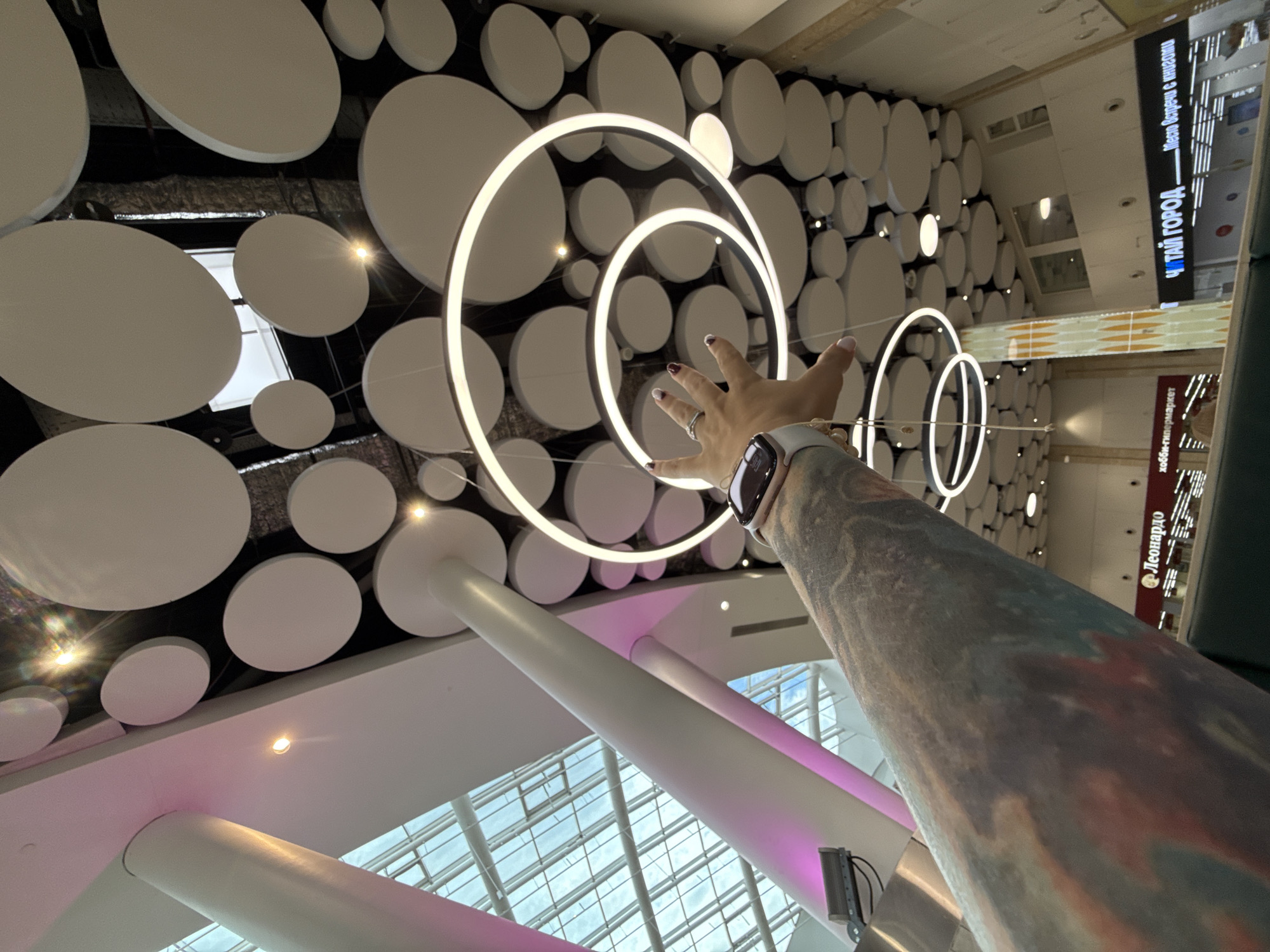 An interior shot is taken from a low angle looking up towards a ceiling adorned with numerous white circular acoustic panels and modern ring-shaped light fixtures. The scene appears to be inside a large commercial space, possibly a mall or atrium. A person's tattooed arm and hand are prominently featured in the foreground, reaching upwards towards the lights. The hand has manicured nails with red polish and wears a smart watch on the wrist. In the background, visible through a glass ceiling or skylight, is a cloudy sky. Lower down, signage for "Леонардо" and "ЧИТАЙ ГОРОД" are visible, indicating retail stores. The lighting is a mix of ambient light from the ceiling fixtures and natural light filtering through the glass. The overall impression is one of contemporary interior architecture with a focus on design elements.