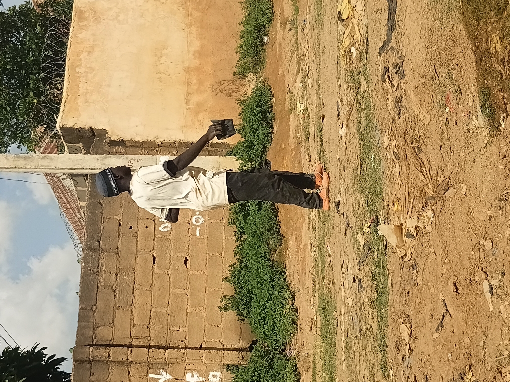 A young Black male, wearing a white t-shirt, black pants, a dark blue cap, and orange shoes, stands facing right in what appears to be a dusty, unpaved street in Jos, Nigeria. He is holding a black object in his right hand. Behind him, a brick wall stretches across the scene, topped with barbed wire. Green bushes grow along the base of the wall, and to the right, a cream-colored building with a rough texture is visible. The ground is littered with debris, including plastic bags, dry grass, and other trash. The sky above is partly cloudy. The overall impression is one of an outdoor urban environment in a developing area.