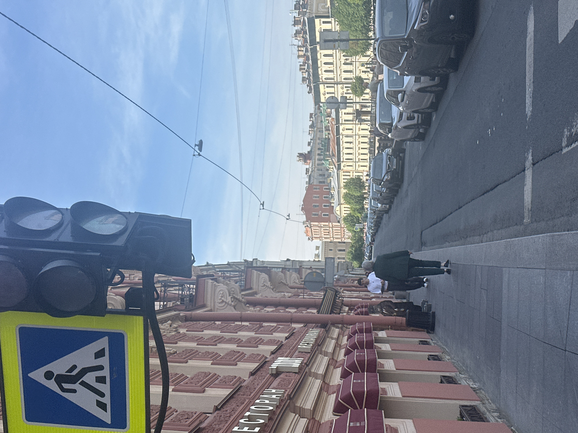 An outdoor, eye-level, medium shot captures a city street scene under a clear blue sky. A pedestrian crossing sign is prominently displayed on the left, alongside a traffic light. The street features a sidewalk with a woman in a dark green jacket walking away from the camera, and a man in a white shirt standing. Buildings with red and cream facades line the street, many featuring awnings. A row of parked cars is visible along the street, with apartment buildings in the background.