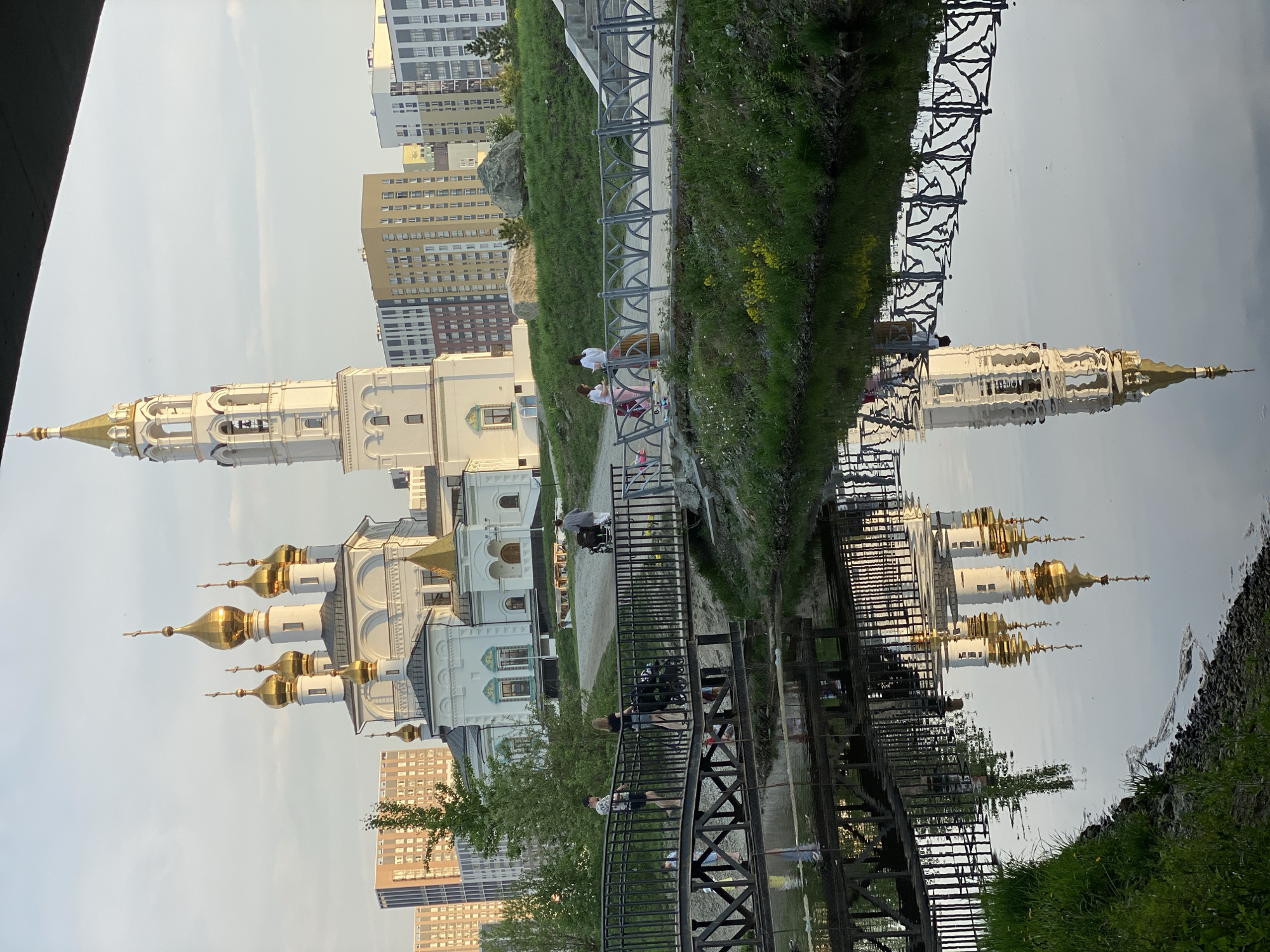The image depicts a scene in Yekaterinburg, Russia, reflected in water, with a church and modern buildings in the background. The church, featuring white walls and golden domes, stands next to a cluster of high-rise apartment buildings. A pedestrian bridge crosses the water, and several individuals are visible on or near the bridge. One person is in a wheelchair. Lush green grass grows on the bank. The sky is bright.