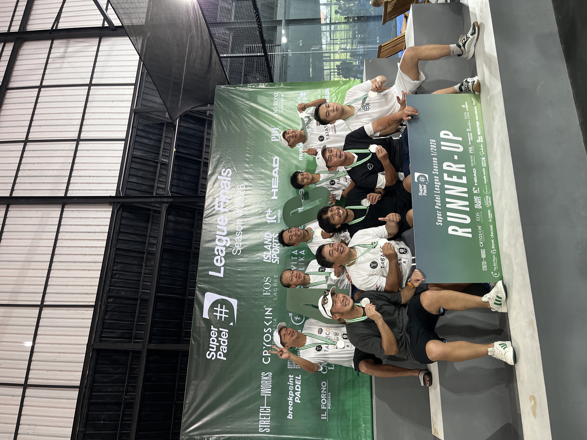 This indoor shot, taken in North Kuta, Indonesia, features a group of men posing for a photo with a "Runner-Up" banner for the Super Padel League Season 1/2026. The men are wearing medals and sports attire with the "Tabasco" logo, and they are positioned in front of a large advertisement promoting the event, with sponsors listed, including "EOS ISLAND SPORTS", "BREAKPOINT PADEL", and "IL FORNO PIZZERIA". Some of the men are holding a sign that says "RUNNER-UP". The background reveals parts of the building's structure, including the ceiling and wall.