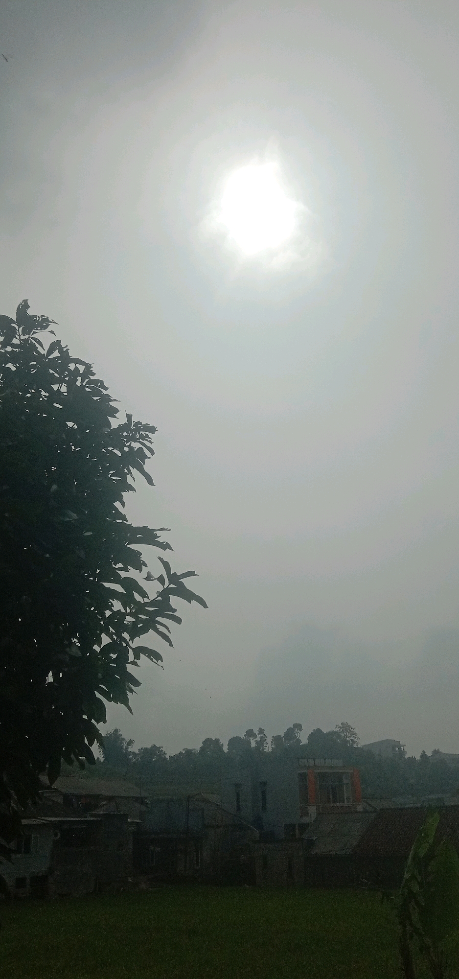 The image depicts a hazy sky with the sun visible through a layer of clouds. In the foreground, to the left, is a dark silhouette of tree branches with leaves. Below the sky and tree, a village scene unfolds, featuring a cluster of buildings with varied rooflines and architectural styles. Some structures appear to be residential homes, while others may be commercial or utility buildings. The buildings are situated on a slight incline, with a green grassy field in the lower portion of the image. In the background, a dense line of trees forms a natural barrier against the sky, suggesting a rural or semi-rural setting. The overall atmosphere is muted due to the hazy conditions. The location is identified as Sukanagalih, Indonesia. There are no people visible in the image.