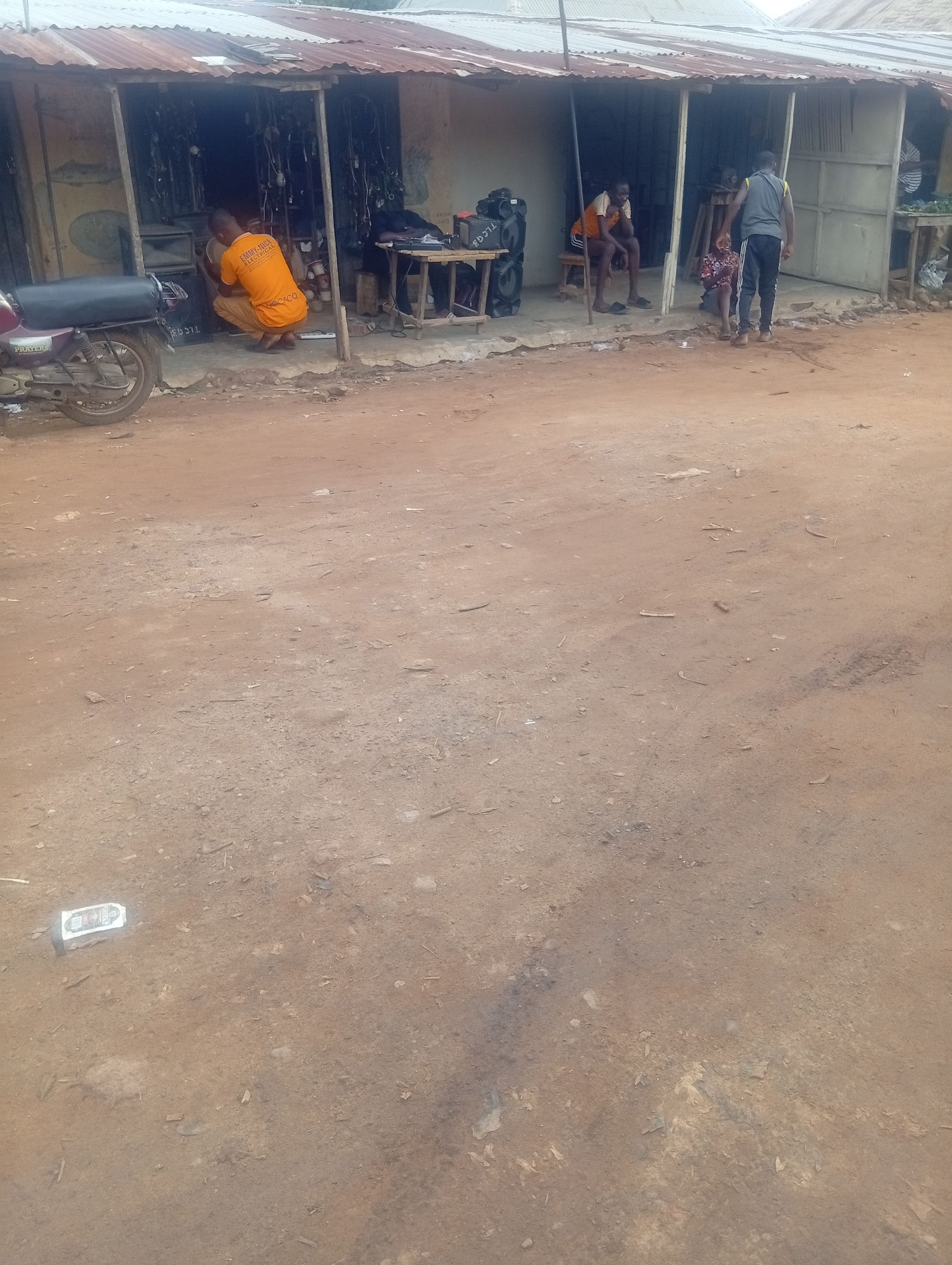 In Yelwa, Nigeria, a street scene unfolds with a small shop in the background. The shop, constructed with corrugated metal roofing and wooden supports, houses various items, including what appears to be electrical wiring. In front of the shop, a person wearing an orange shirt and pants is kneeling, possibly working on something. To the right, another person in athletic wear is standing, facing away from the camera. A child in a patterned dress is also present near the shop. A motorcycle is parked to the left of the shop. The ground in the foreground is unpaved, with loose soil and scattered debris. A white object, possibly a discarded can, lies on the ground to the lower left. The overall environment suggests a bustling local marketplace or a roadside business area.