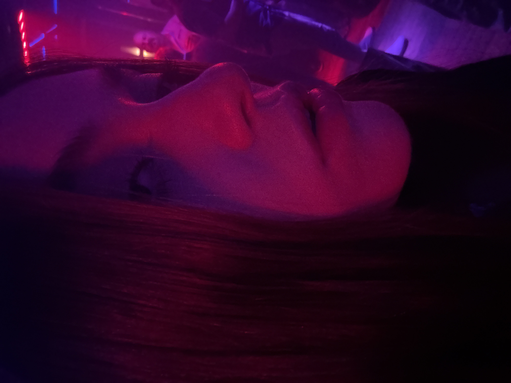 A close-up, low-angle shot depicts a person's face in profile, illuminated by red and purple light. The person is lying down on a surface of dark brown hair. The background is dark and blurry, with indistinct objects and possibly other people. The scene is likely indoors due to the artificial lighting and controlled environment. There are no obvious clues that indicate the location to be Mexico City.