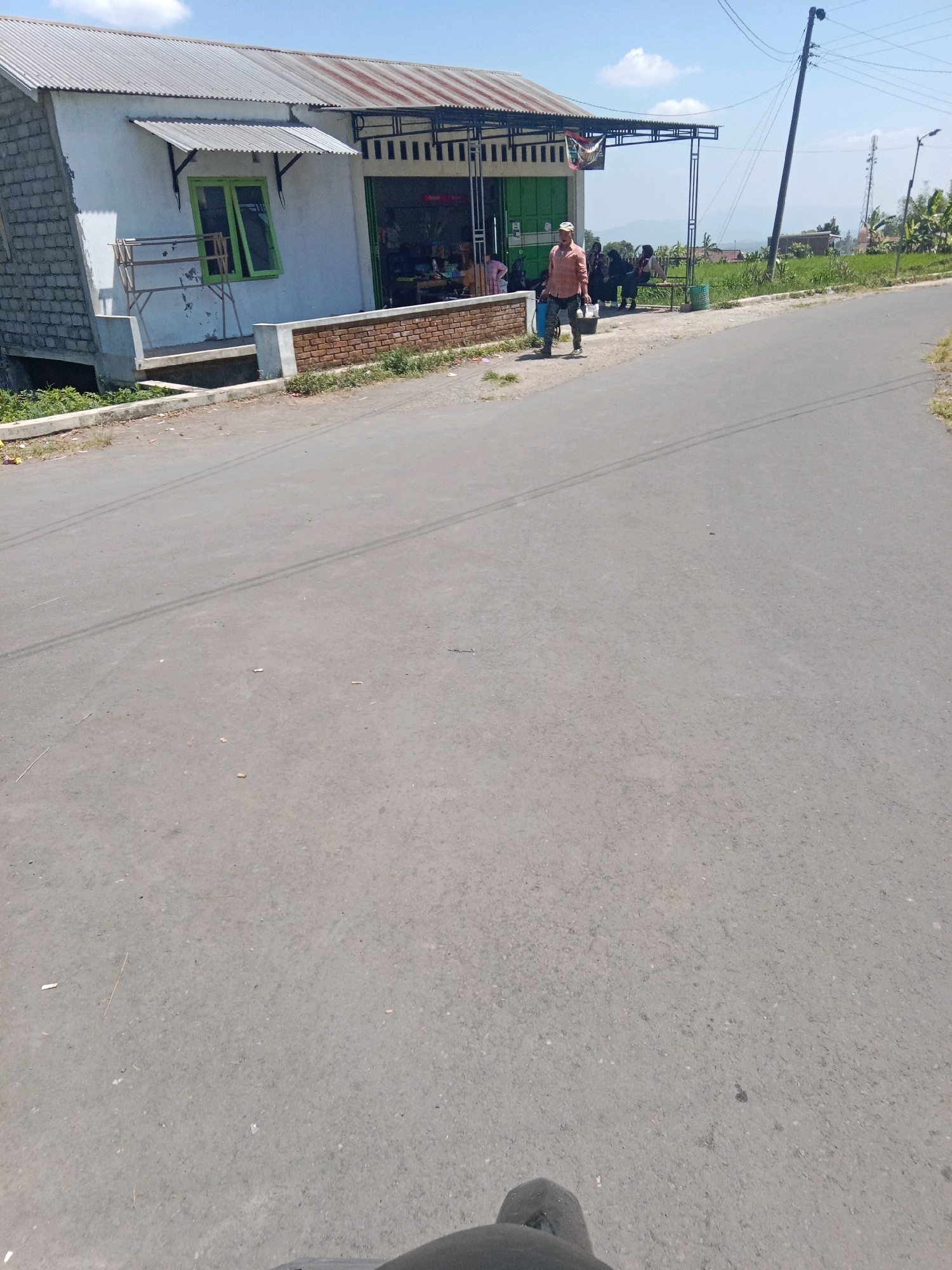 The image depicts a roadside scene in Sumberdalem, Indonesia. In the foreground, a gray asphalt road runs diagonally from the bottom left to the top right. A person, wearing a light orange long-sleeved shirt and dark pants, walks along the side of the road, carrying a white bucket in their right hand and a dark bag over their left shoulder. They are walking away from a small building with a corrugated metal roof and white walls, which appears to be a shop or residence. The building has a green-framed window and an awning extending over the front. Several other individuals are seated near the building, appearing to be engaged in conversation. Behind the building, lush green fields and vegetation are visible, with hills in the distance. A utility pole with wires stands to the right of the road, and a communication tower is visible further in the background. The sky is clear and blue with a few scattered clouds.