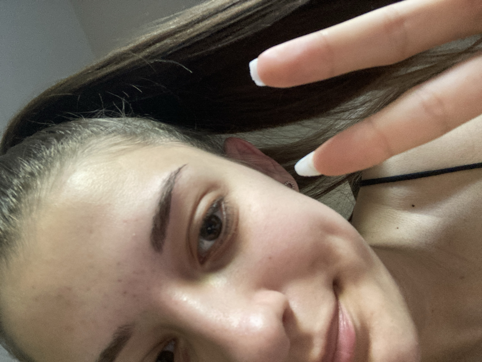 A close-up, selfie-style shot of a light-skinned individual with brown hair. The person is making a peace sign with their right hand, the fingers prominently displayed with white-tipped nails. The individual's face is partially visible, showing a brown eye, eyebrow, cheek, and part of the nose and lips. The background is out of focus, but a dark strap is visible near the neck, and a mole is present on the shoulder area. The location is not discernible from the image.